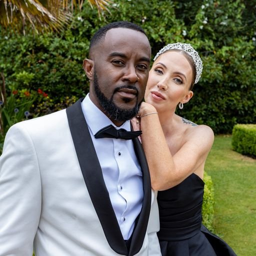 A couple dressed in formal attire posing outdoors. The man is wearing a white tuxedo jacket with a black bow tie, and the woman is in a black dress and tiara, wrapping her arm around him. They are standing in a garden setting with greenery in the bac