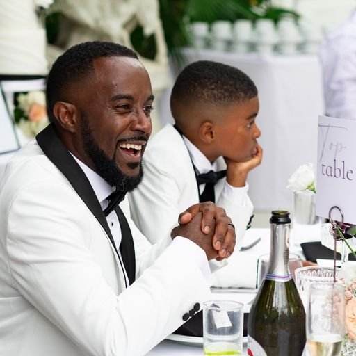 A man in a white suit with a black tie smiling at a table next to a young boy in similar attire at a formal event. The table is set with a wine bottle, glassware, and a "top table" sign.