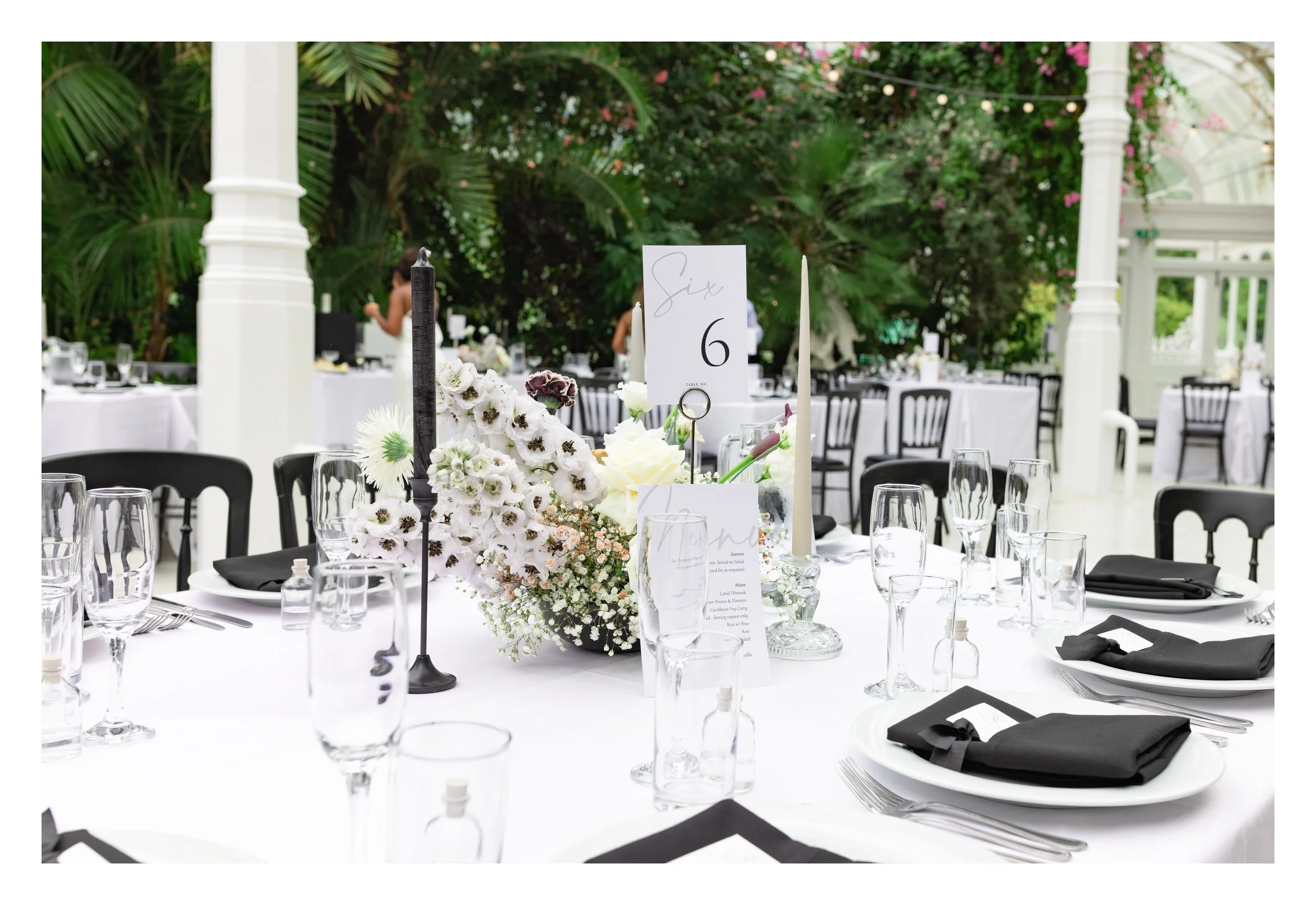 Elegant wedding reception table setup with white tablecloth, black chairs, black napkins, floral centerpiece, glassware, and table number six in a garden setting.