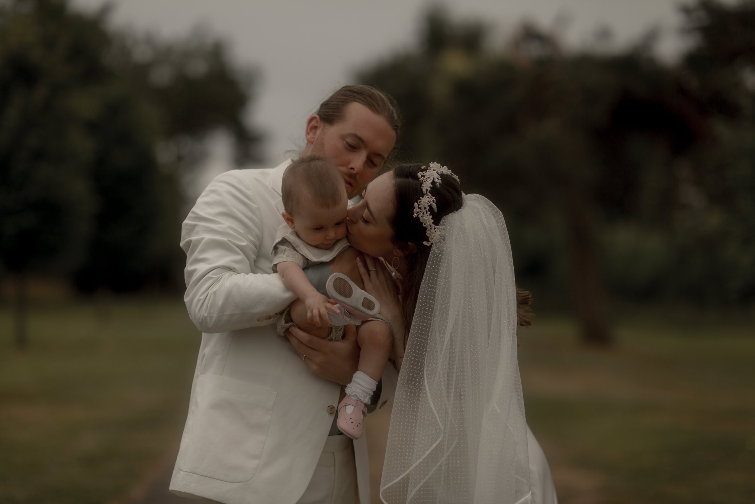 Wedding couple holding a baby outdoors, with the woman in a veil and floral headpiece