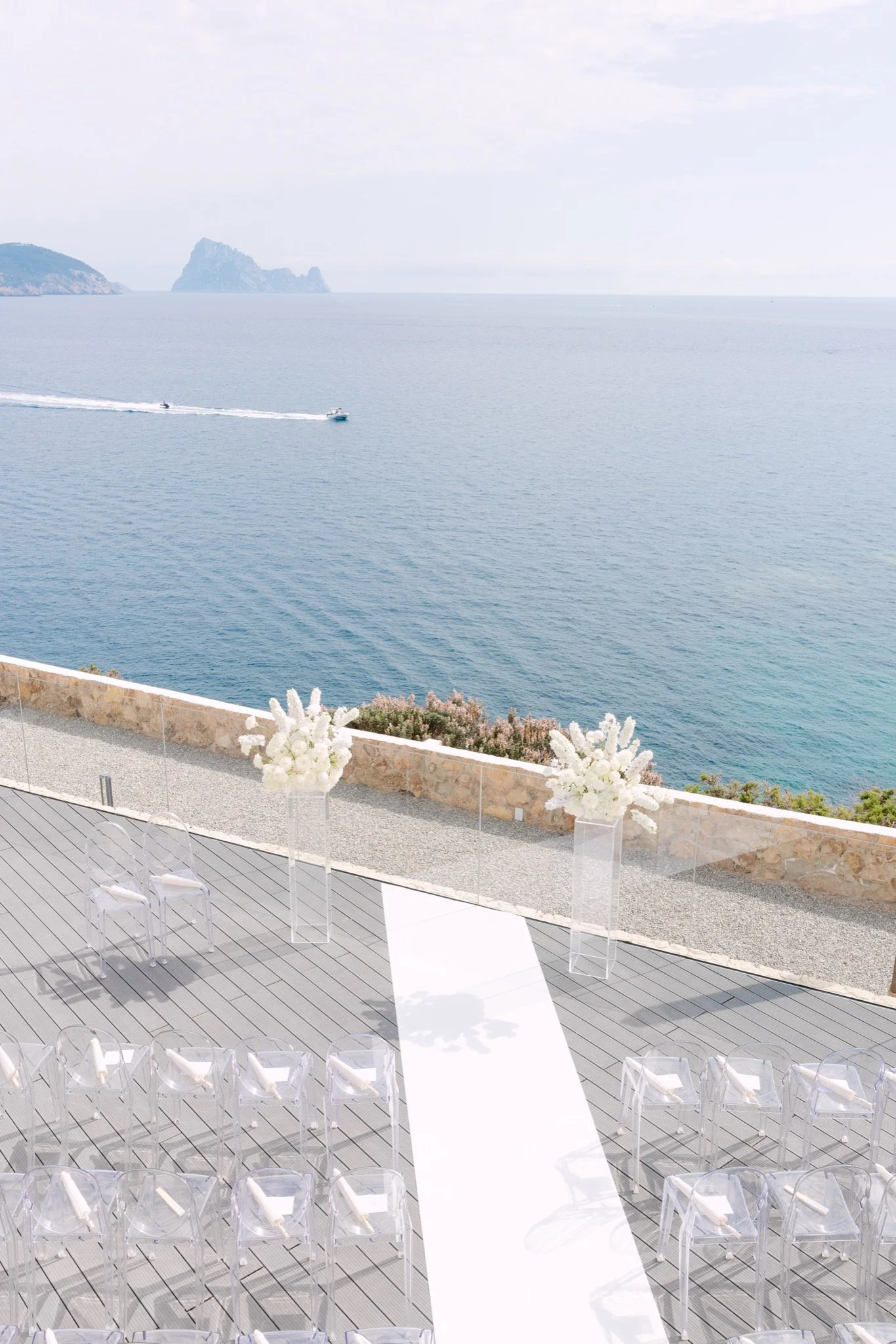 Luxury cliffside wedding ceremony setup at 7Pines Resort Ibiza overlooking Es Vedrà, featuring clear ghost chairs and white floral columns by Flowers Ibiza, planned by Stacey Koks Weddings.