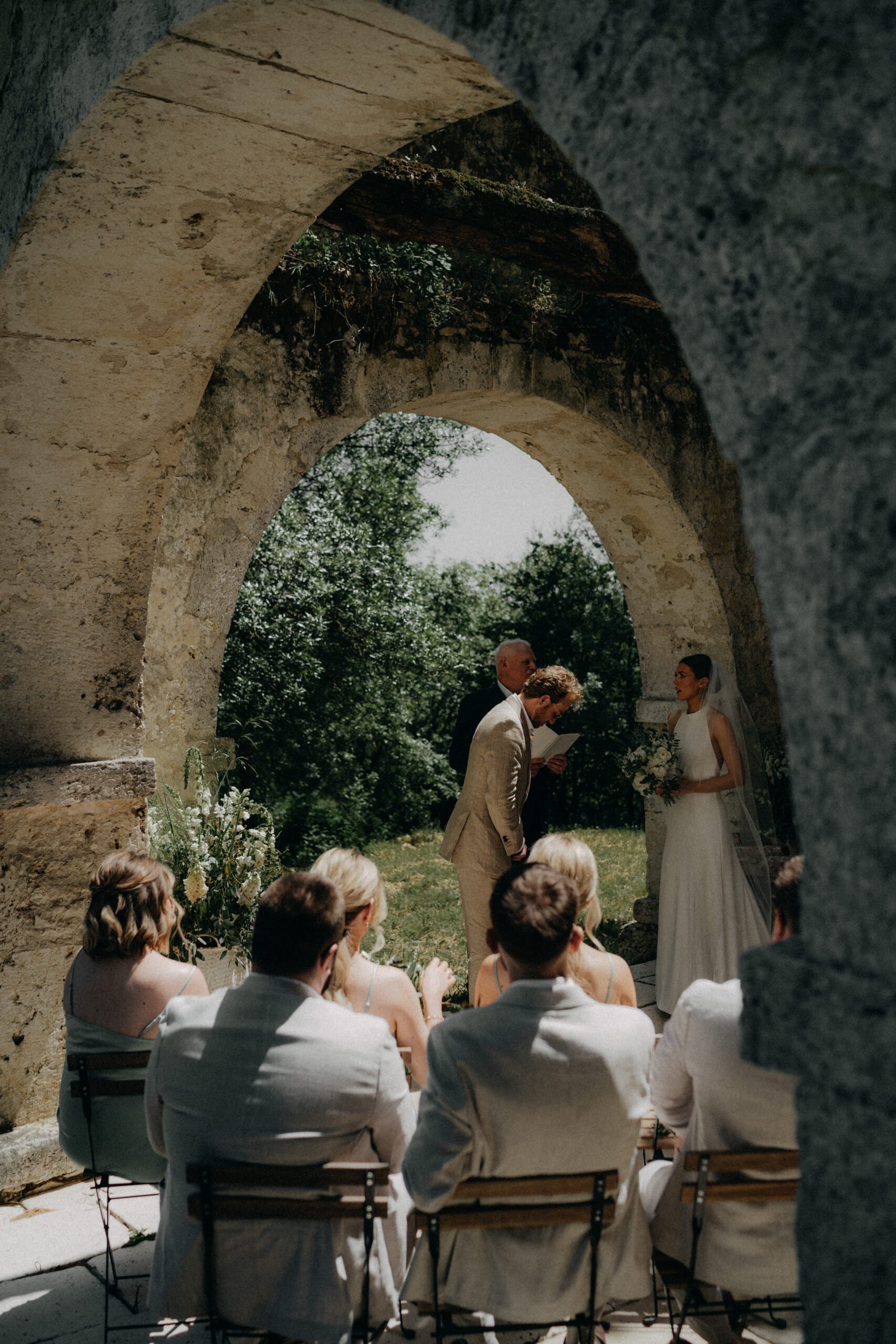 Outdoor wedding ceremony under an ancient stone archway with a bride, groom, officiant, and seated guests.