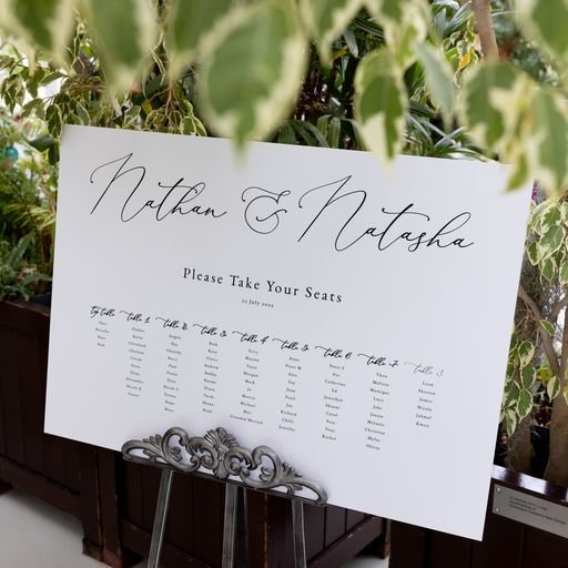 Wedding seating chart with elegant calligraphy, titled "Nathan & Natasha." A list of table assignments is displayed on a white board, surrounded by plants.