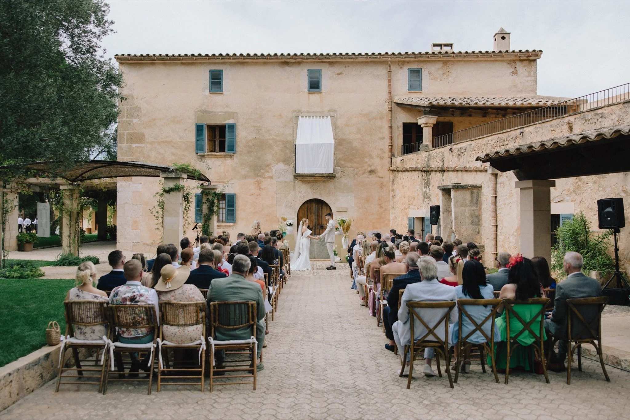 Outdoor destination wedding ceremony in a historic stone courtyard with guests seated on wooden chairs, planned by Stacey Koks Weddings in Mallorca.
