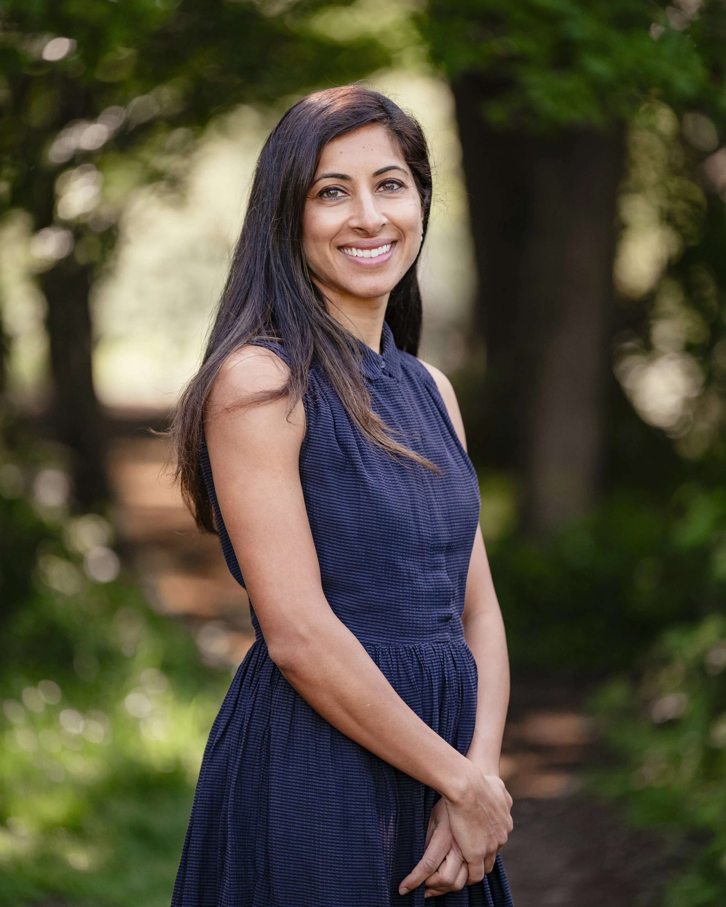 A woman with long dark hair and light skin smiling outdoors, wearing a navy blue sleeveless dress.