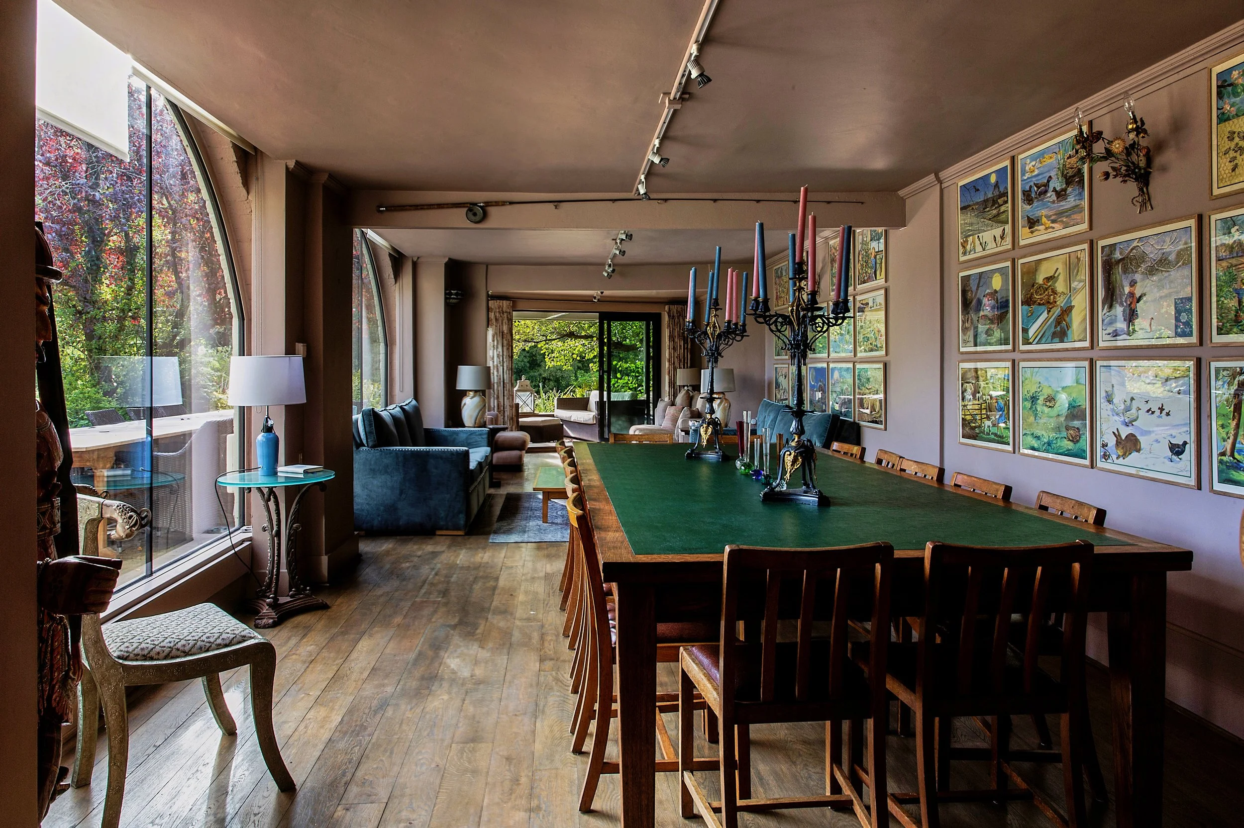 Elegant dining room with large windows, wooden flooring, and a long table with a green felt surface, adorned with colorful candles. The room features wall art, floor lamps, and a cozy seating area with sofas and chairs, opening to a garden outside.