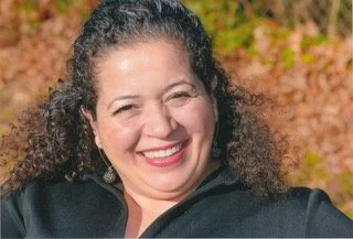 A woman with curly hair smiling outdoors during autumn with colorful fall foliage in the background.