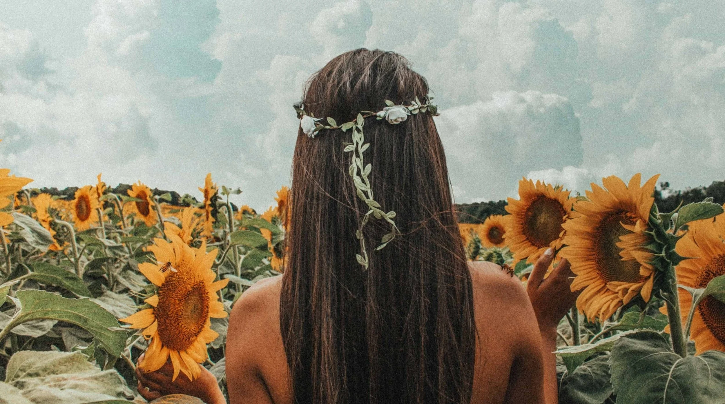 A woman with long brown hair and a floral crown stands in a sunflower field, facing away from the camera, touching a sunflower.
