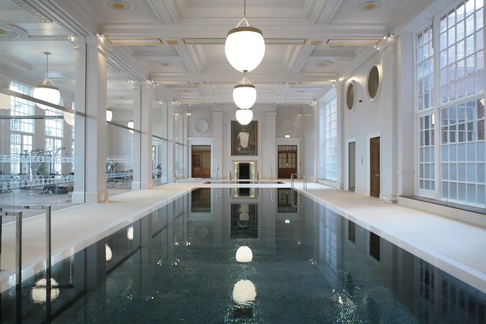 LA London Architects LLP — Indoor swimming pool in a luxurious, well-lit room with large windows, white walls, and elegant chandeliers.