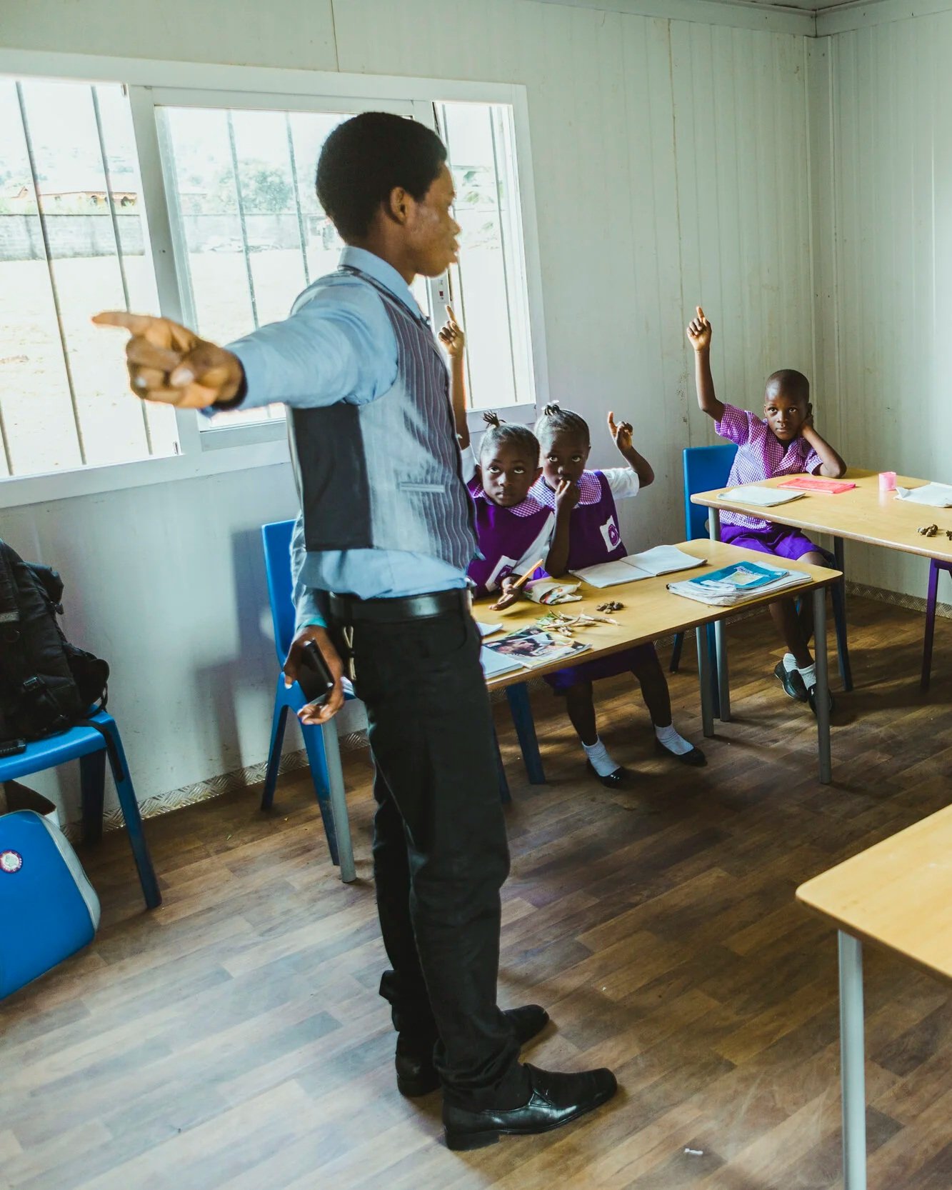 A classroom with young students raising their hands while a teacher stands at the front. The students sit at desks with books and supplies. The teacher is pointing towards the students. The room has large windows letting in natural light.