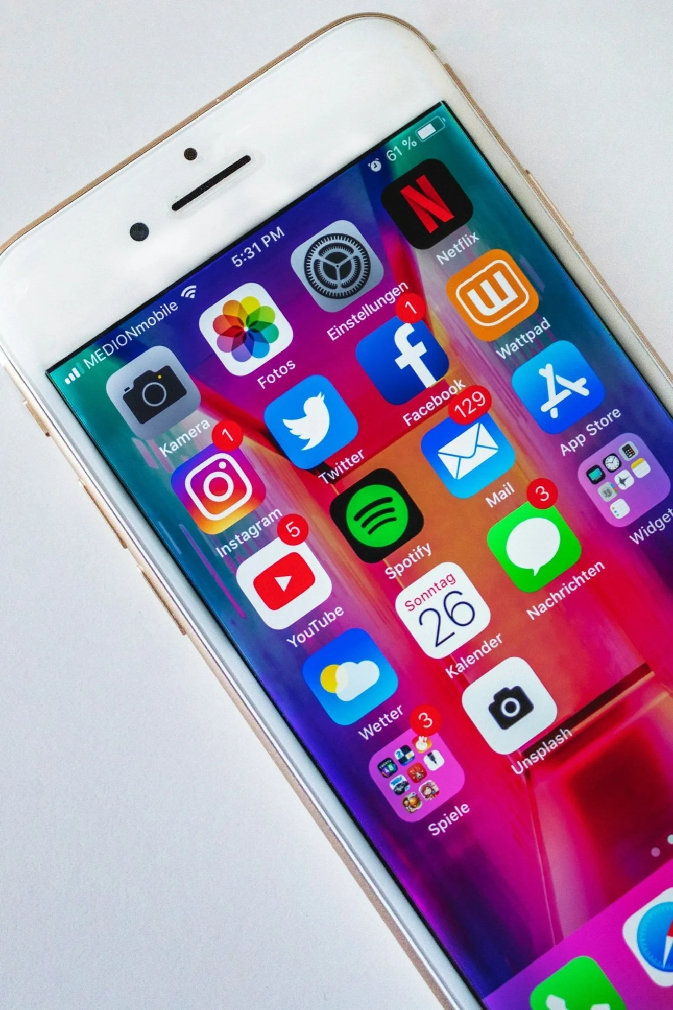 Close-up of an iPhone screen showing social media and app icons on the home screen, with a colorful background. The screen displays various app notifications, including one message from Facebook, YouTube, and Instagram. The phone is placed on a white surface.