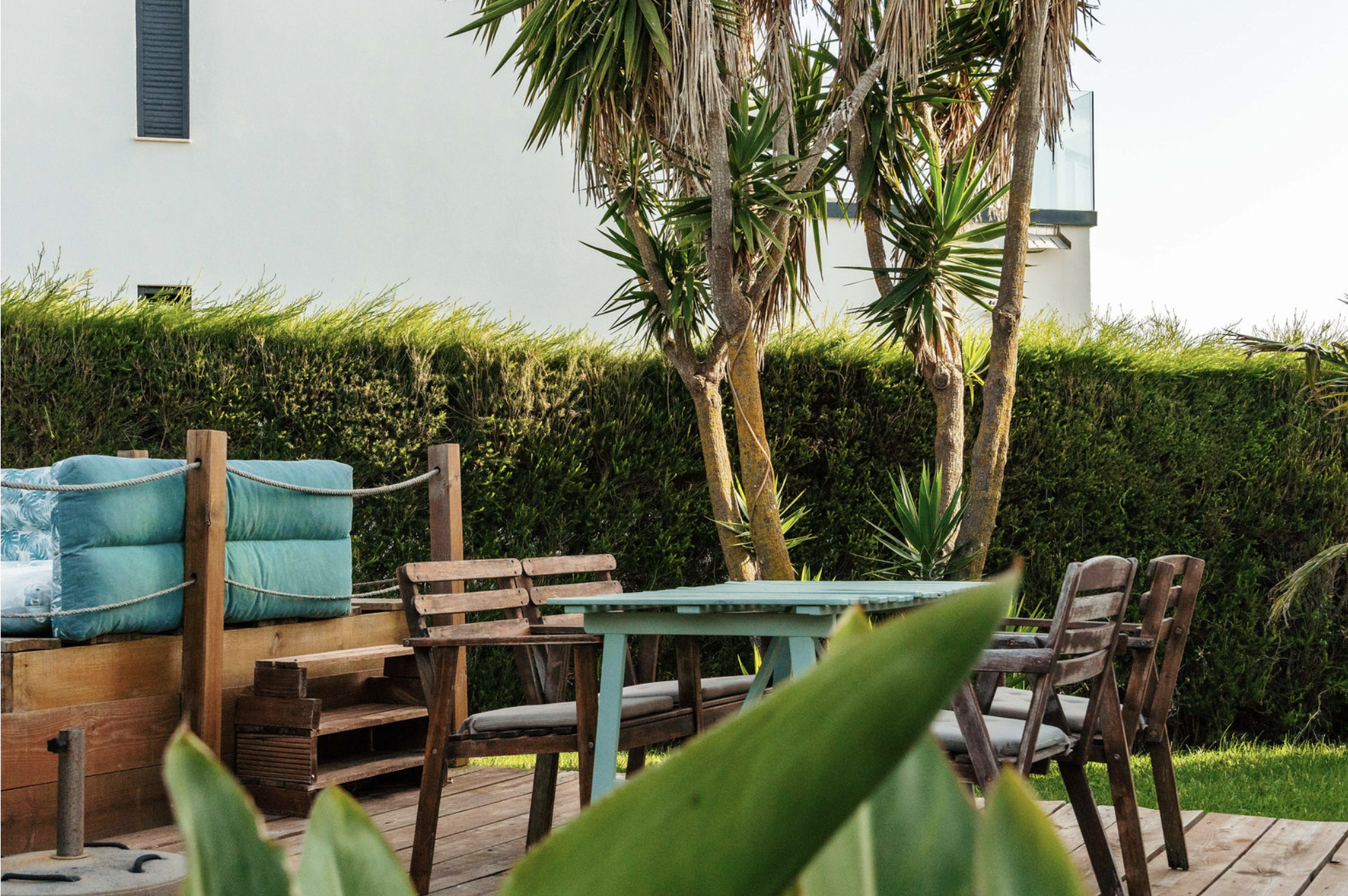 An outdoor patio area with a wooden deck, surrounded by green plants and trees, featuring a turquoise couch with patterned pillows, wooden chairs, a turquoise table, and a background of a modern white building with black shutters.