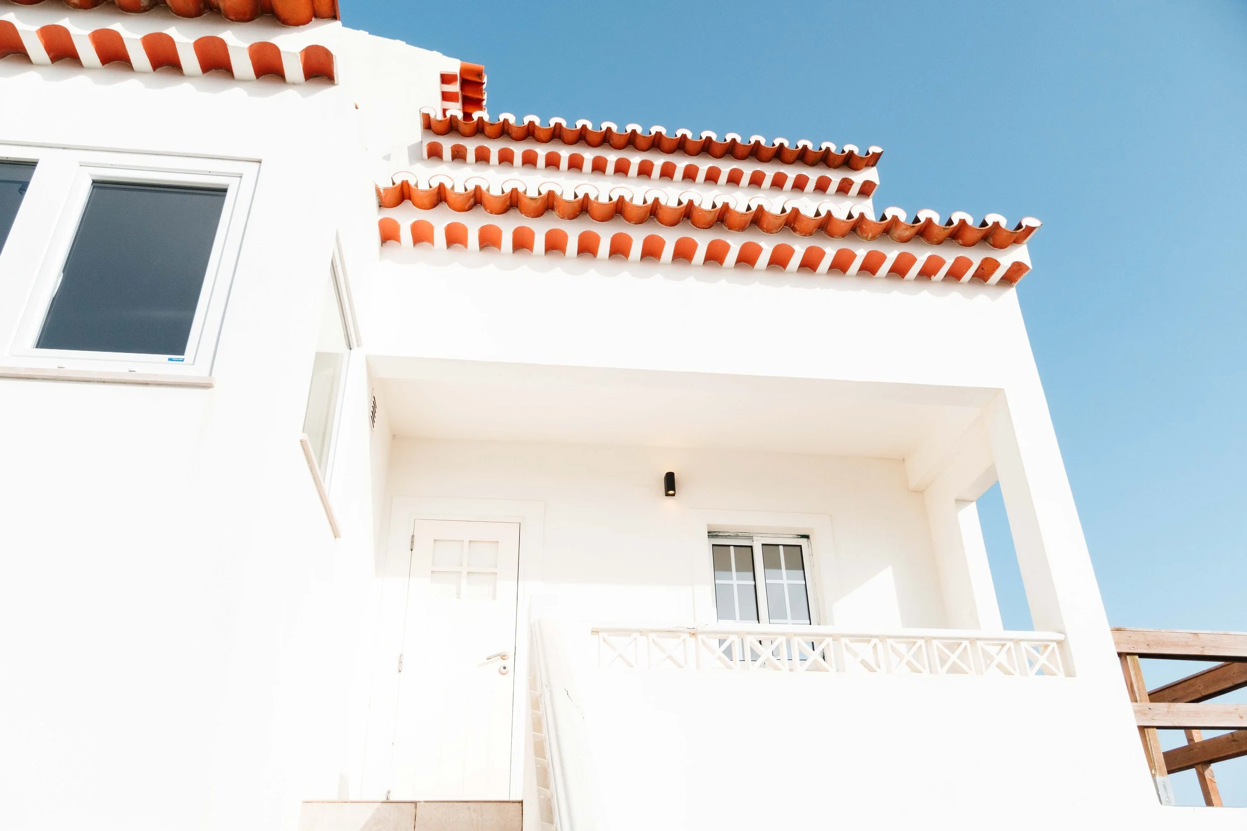 White building with red clay tile roof, window, door, and staircase leading to a balcony, under a clear blue sky.