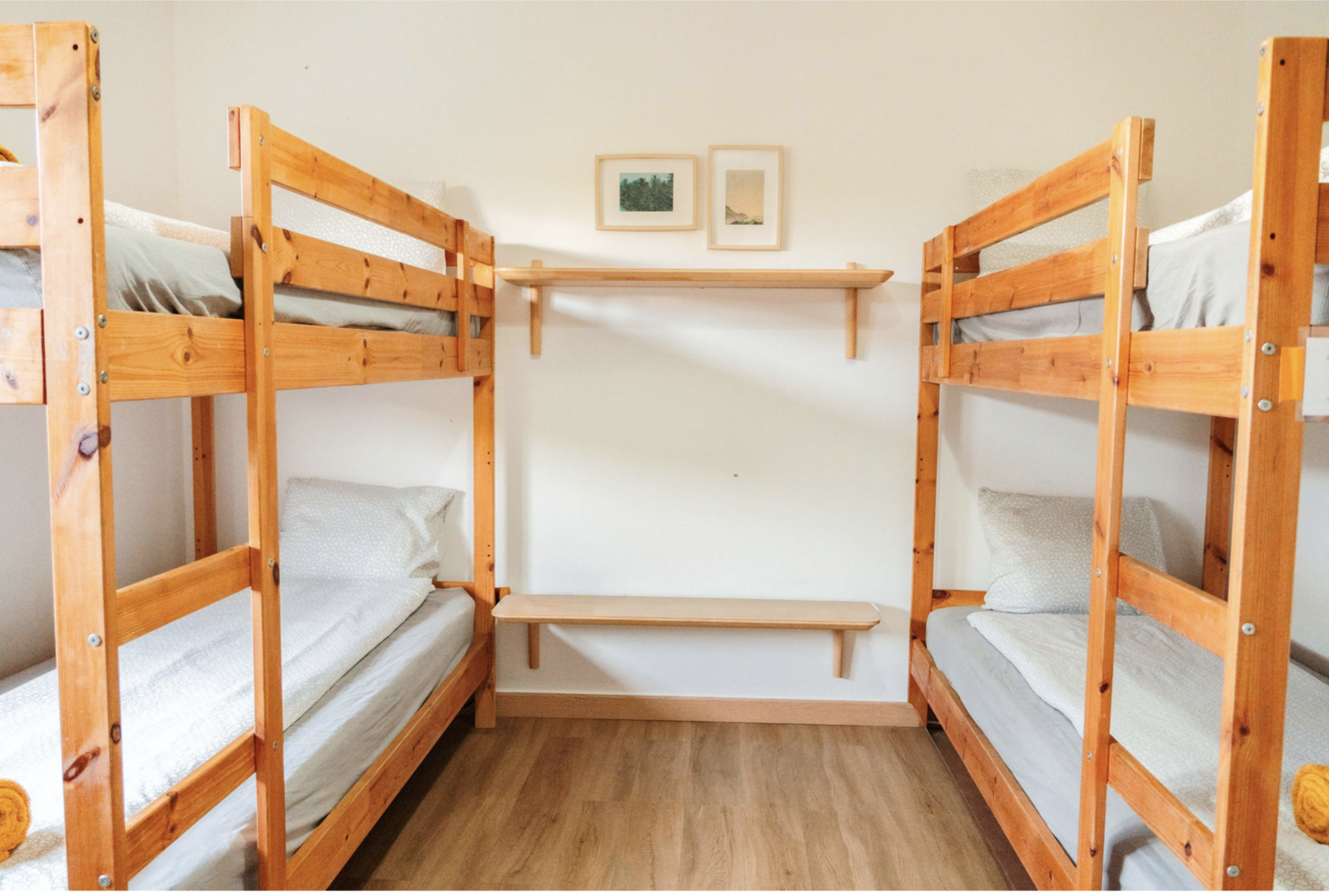 Two sets of wooden bunk beds with white bedding and pillows in a small room, with two framed pictures above a small wooden shelf on the wall.