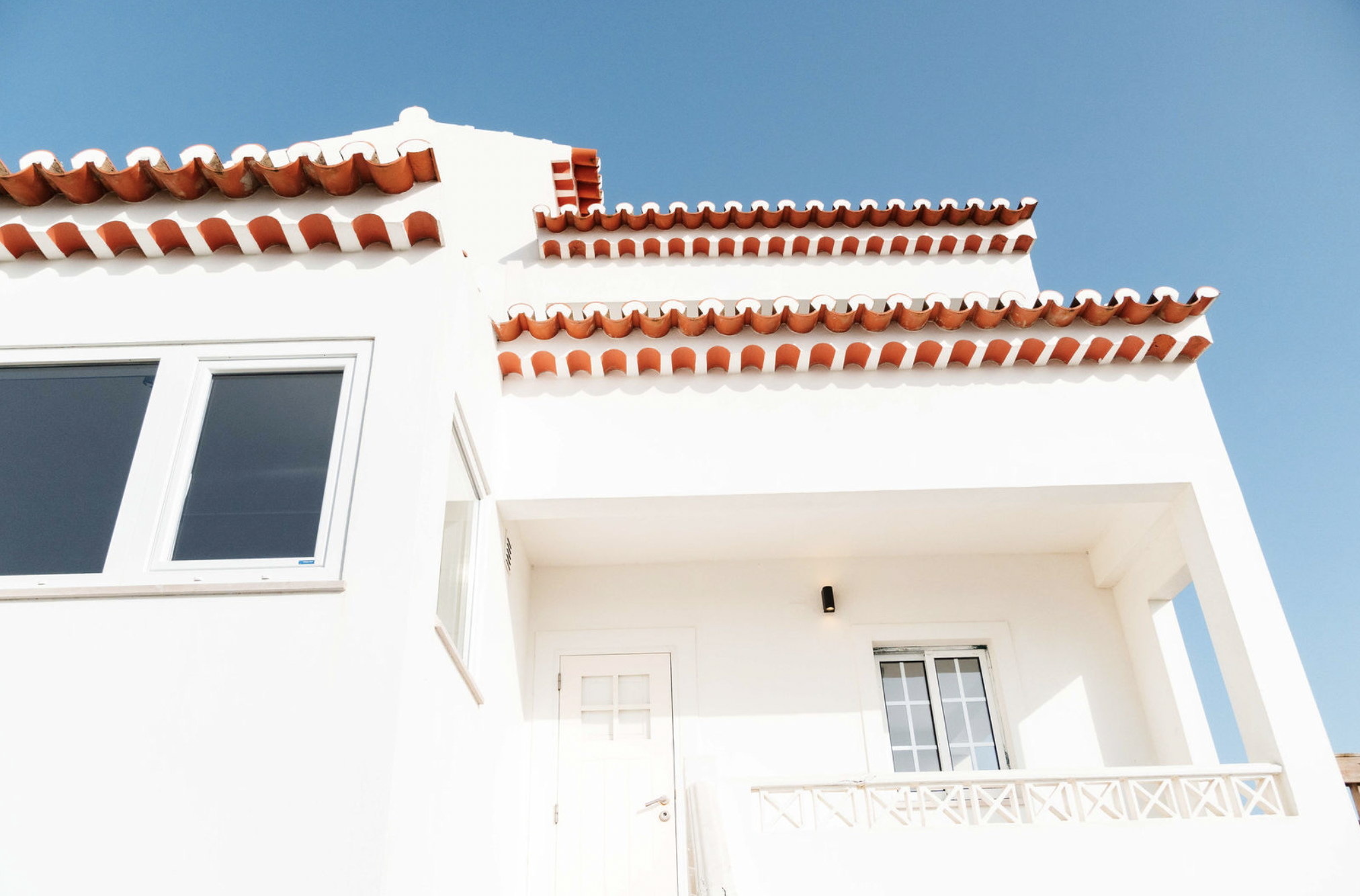White building facade with tiled red roof, windows, and a balcony under a clear blue sky.