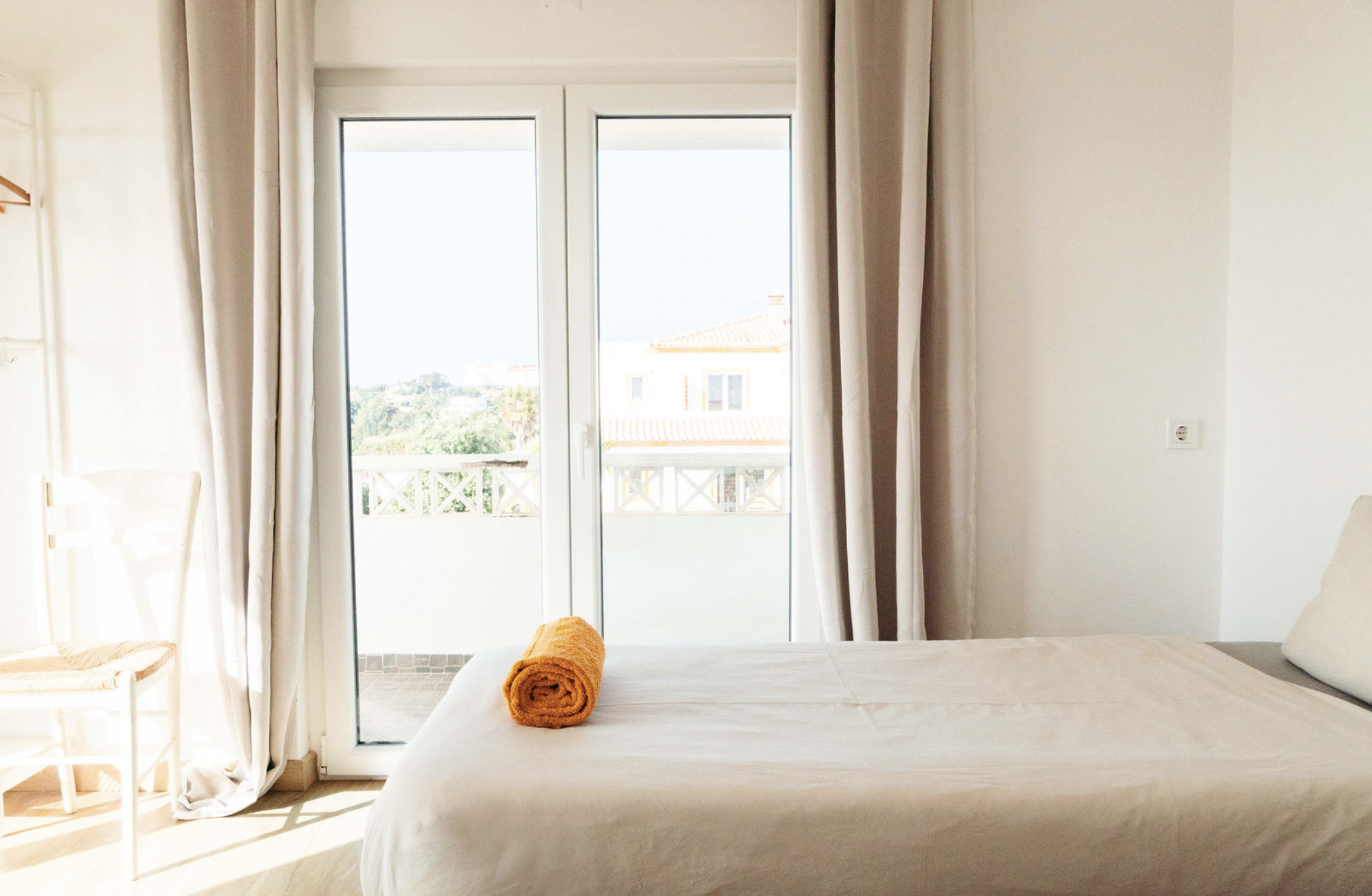 Hotel room with a bed, orange towels, and a balcony with sliding glass door, beige curtains, and a view of neighboring houses and trees.