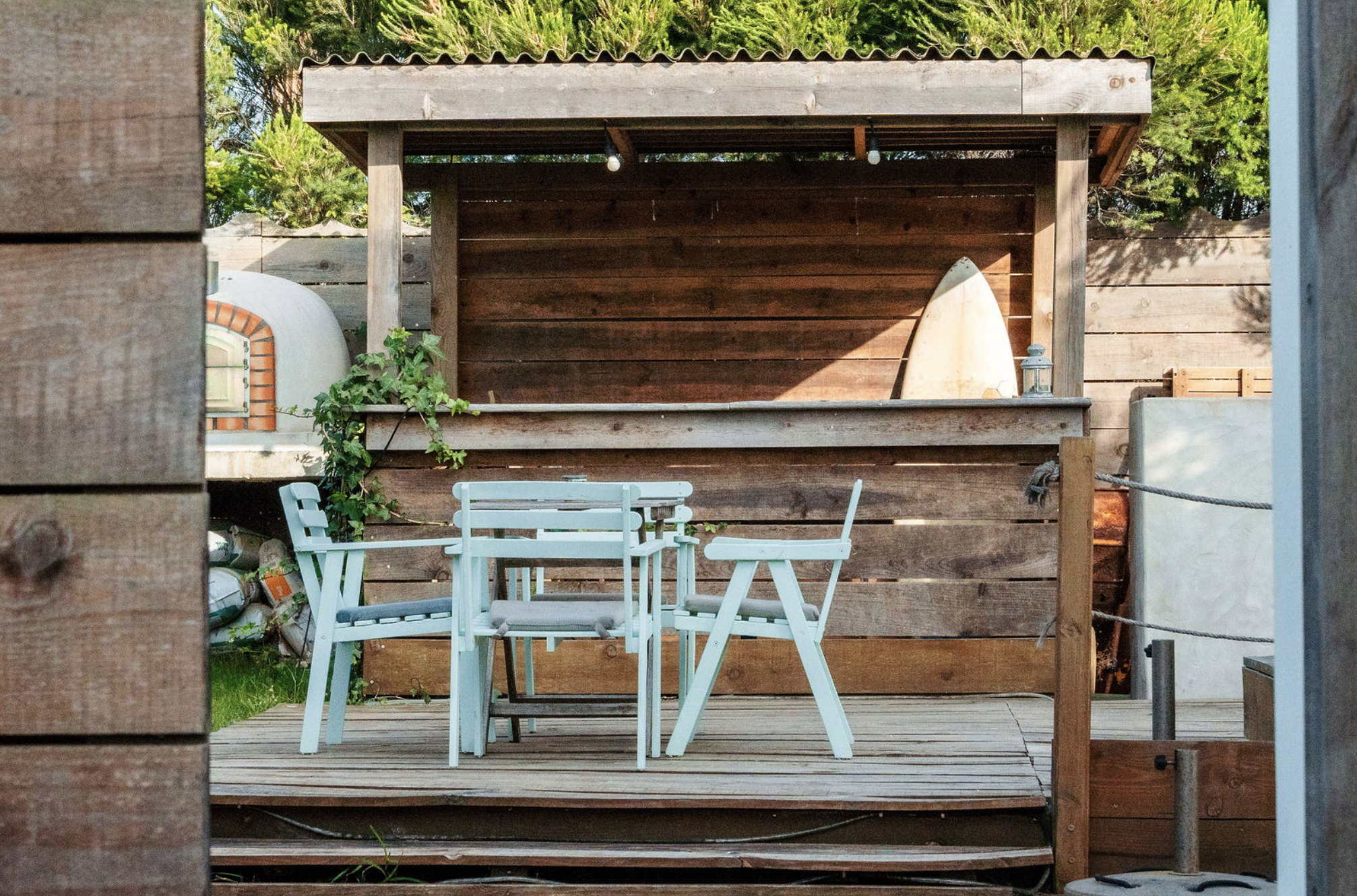 A small outdoor seating area with white chairs and a table on a wooden deck, enclosed by a wooden fence with a wooden shelter overhead. There is a surfboard leaning against the fence and a lantern on the deck. In the background, there are trees and g