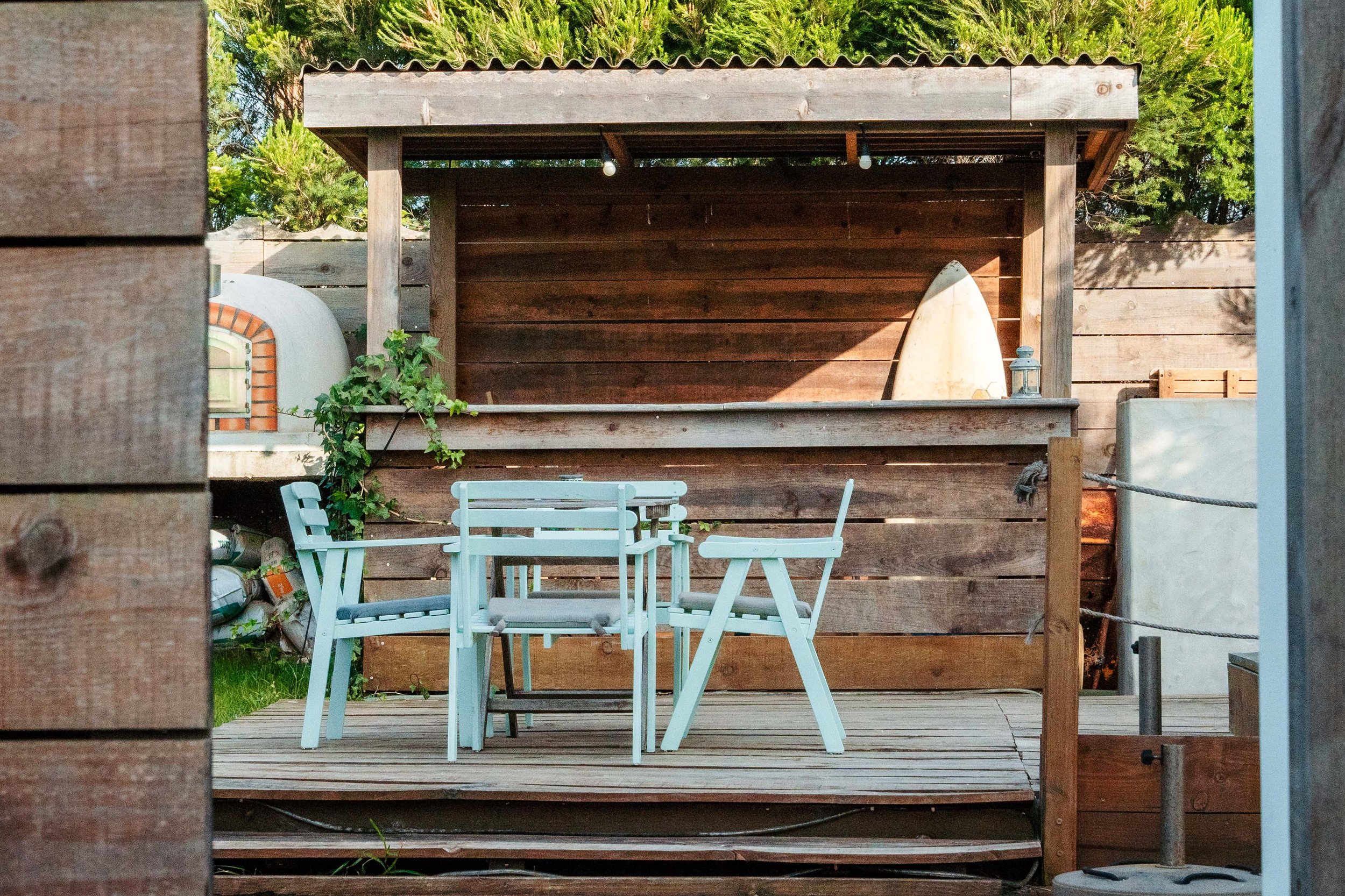 A wooden deck with a white outdoor table and chairs, surrounded by a wooden fence and lush greenery, with a surfboard leaning against the back wall.