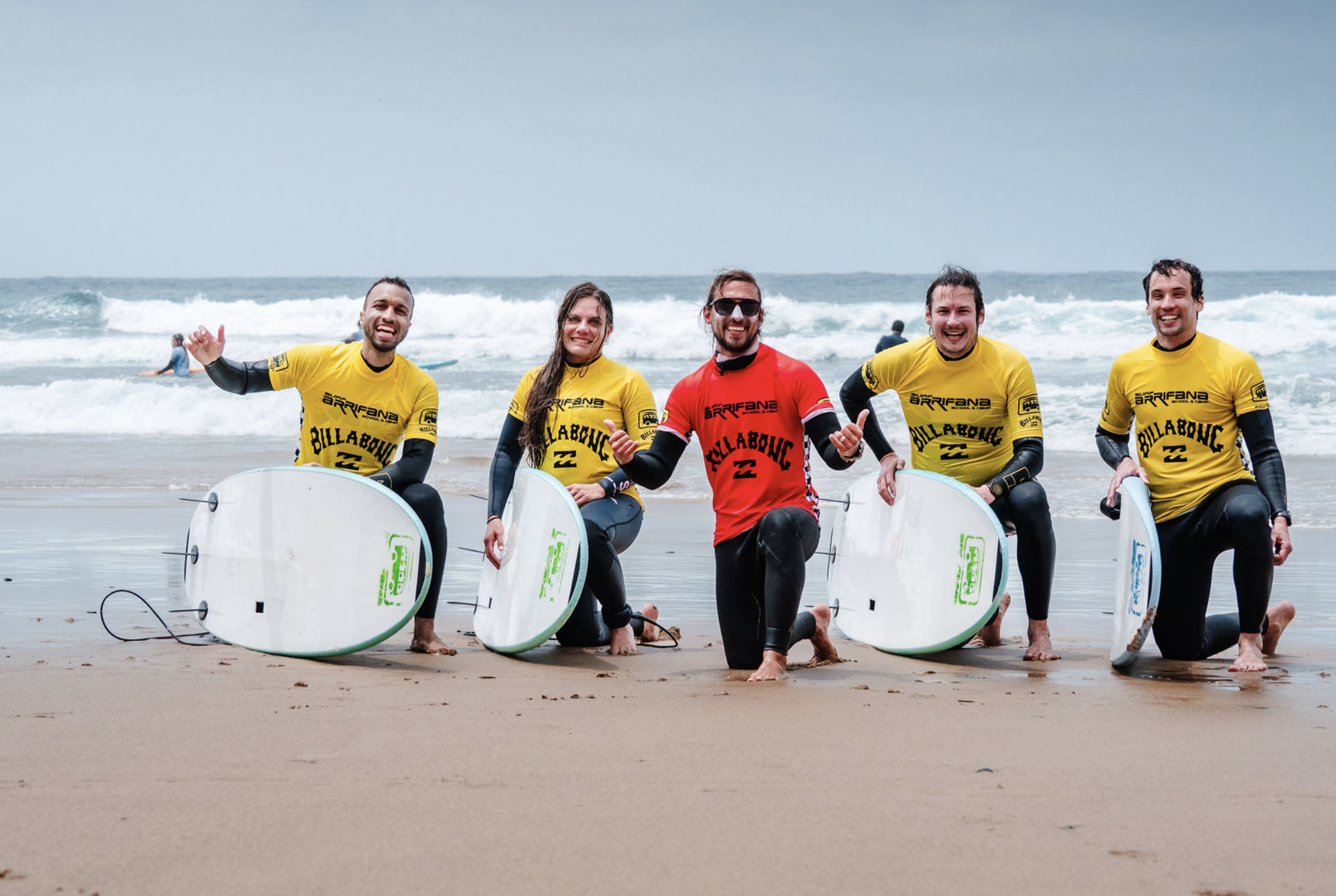 Five people kneeling on the beach holding surfboards, smiling, with waves in the background. Four are wearing yellow Billabong wetsuits, and one person in the center is wearing a red Billabong wetsuit. They appear to be part of a surfing group or tea