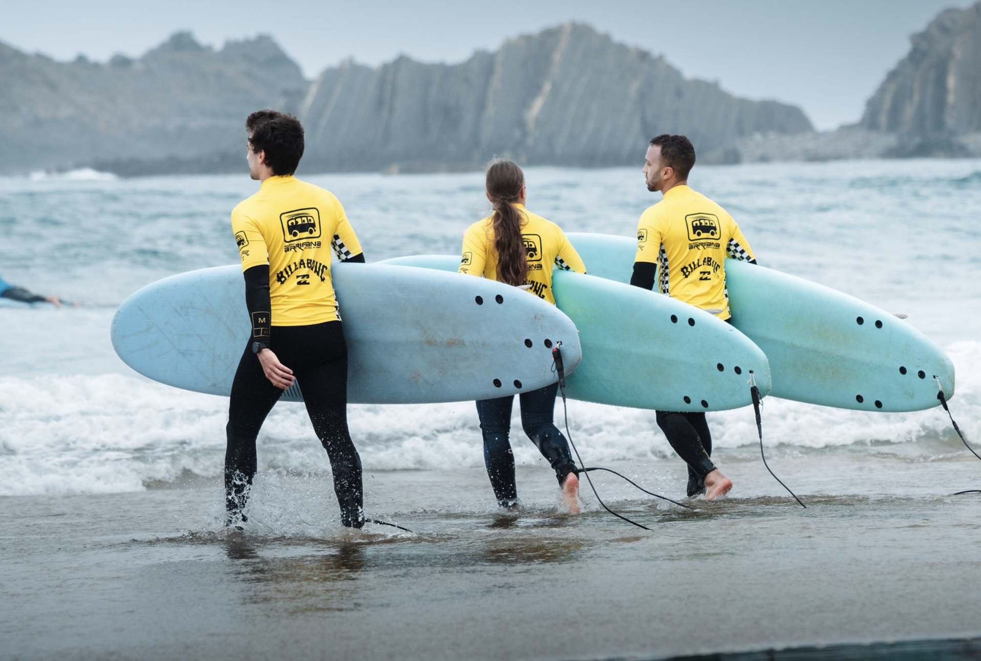Three surfers in yellow rash guards walking out of the ocean carrying surfboards, with rocky cliffs in the background.