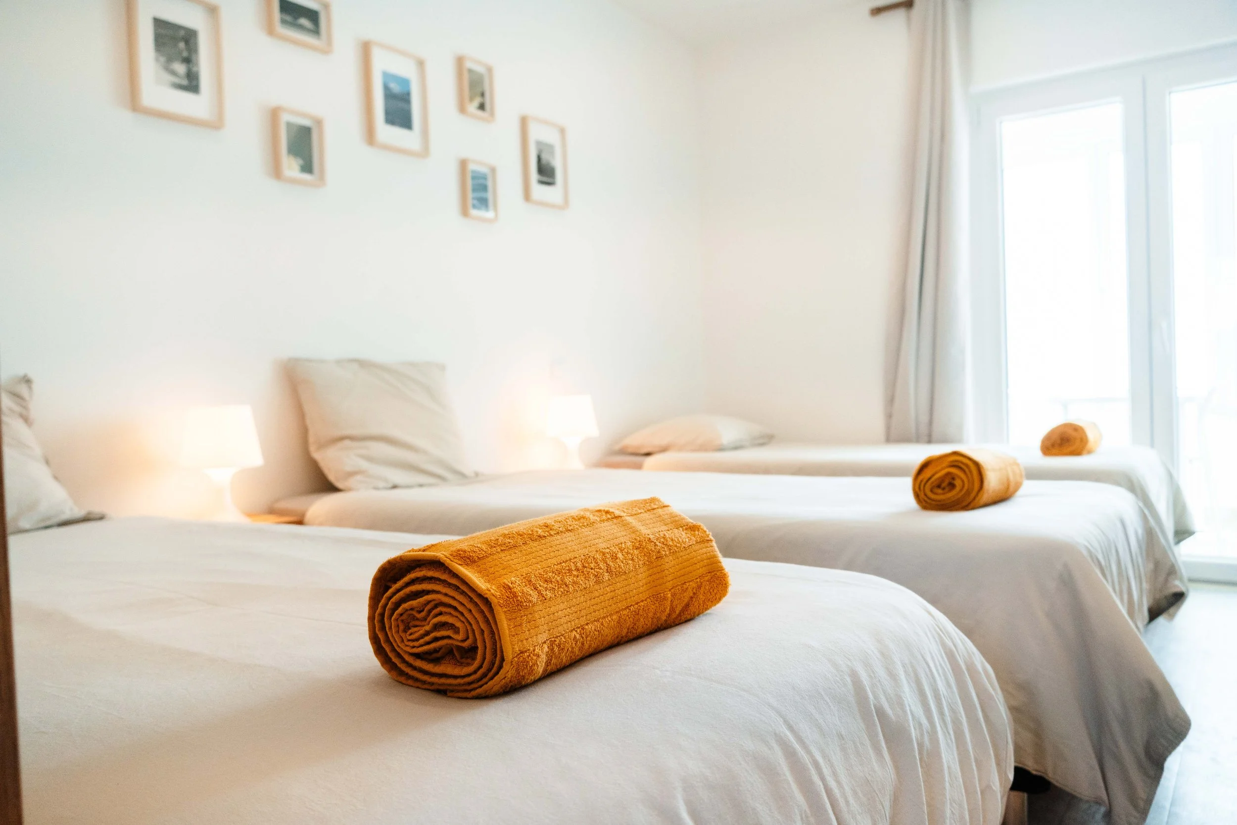 A bright, clean bedroom with three neatly made beds, each topped with a rolled orange towel. The room has many natural light from large windows with white curtains, and a wall decorated with multiple small framed pictures.