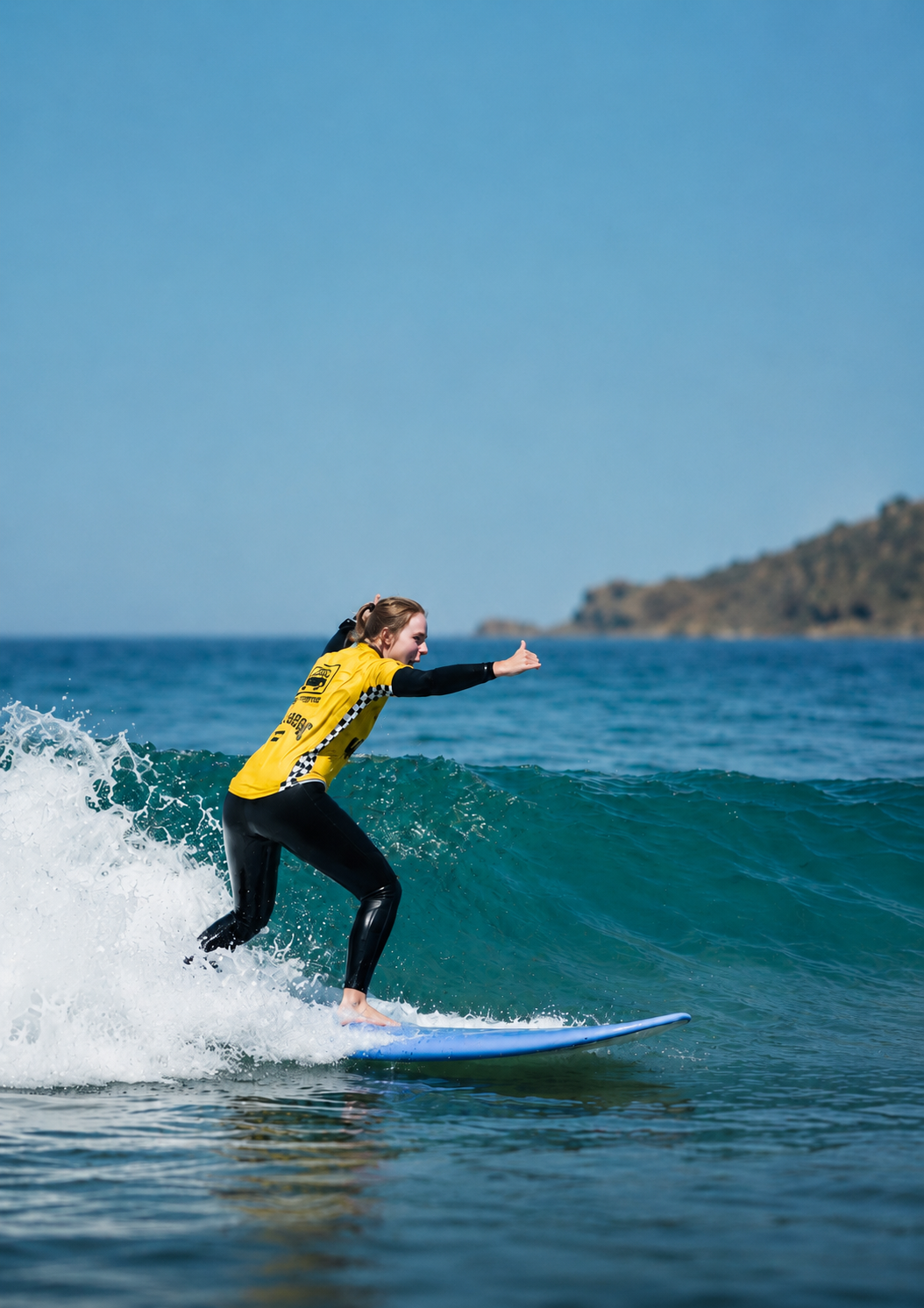 A woman in a yellow and black wetsuit surfing on a blue surfboard in the ocean with a rocky coastline in the background.