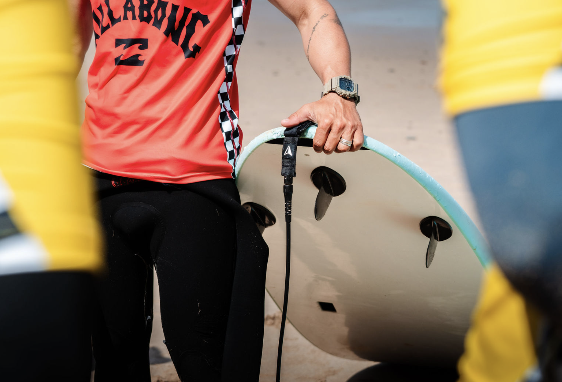 A person holding a surfboard on a sandy beach, wearing a red shirt, a watch, and a ring, with yellow and blue objects around, possibly part of a surfing setup or beach equipment.