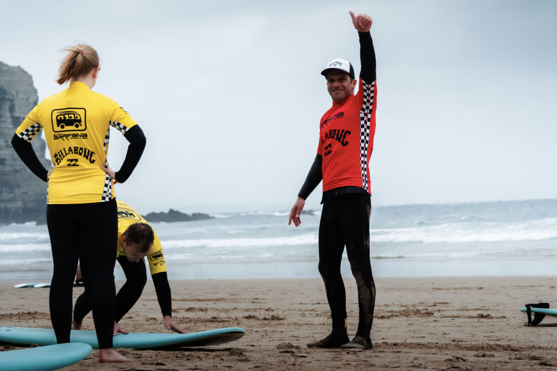 People on a beach preparing for surfing, with one person standing and others bending down near surfboards, wearing wetsuits and yellow shirts with checkered patterns.