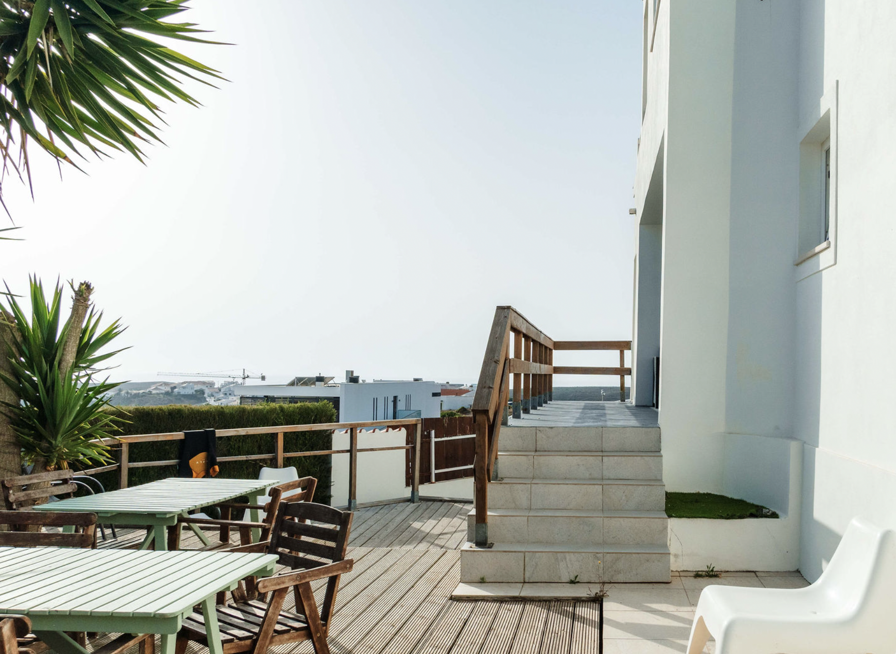 A rooftop patio area with wooden and metal outdoor furniture, potted plants, and a white modern building with small windows. The patio has steps leading to a higher level and a partial view of the surrounding cityscape with buildings and a crane in t