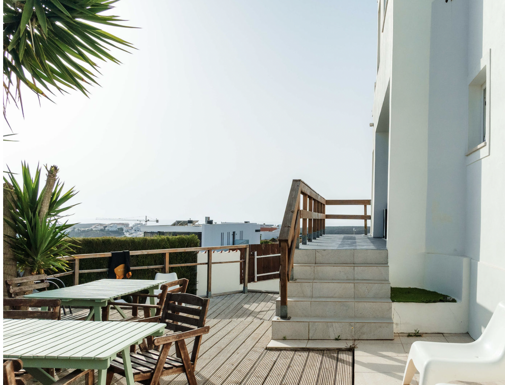 Outdoor patio area with green tables, wooden chairs, potted plants, a white building, and a view of neighboring buildings under a clear sky.