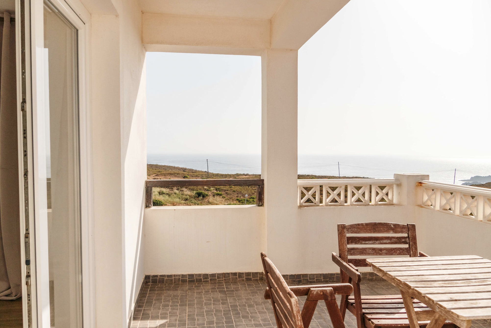 Balcony view with wooden chairs and table, overlooking a landscape with grassy hills and the ocean under an overcast sky.