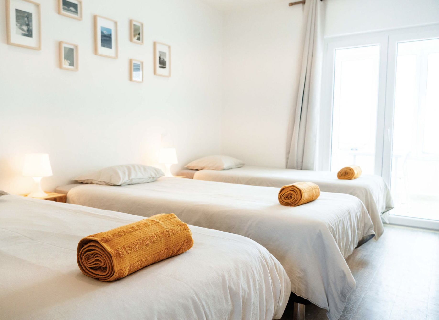 A bright hotel room with three neatly made beds, each with a rolled orange towel at the foot. There are two bedside lamps, and the wall is decorated with several framed photographs. Large windows with white curtains let in natural light.