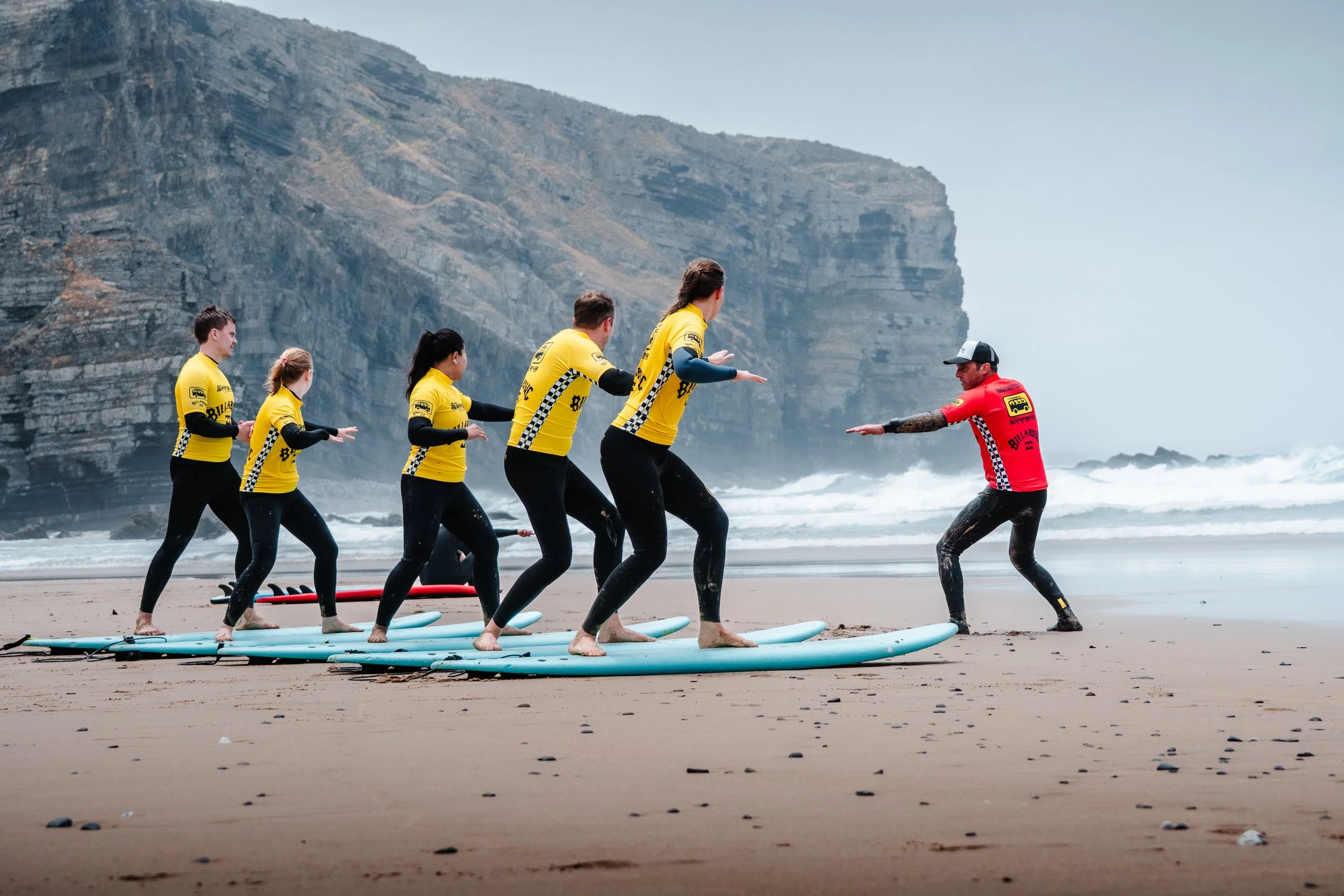 A group of six people in wetsuits and yellow and red shirts, standing in a line with surfboards on the beach, practicing balance exercises while facing each other against a backdrop of cliffs and ocean waves.
