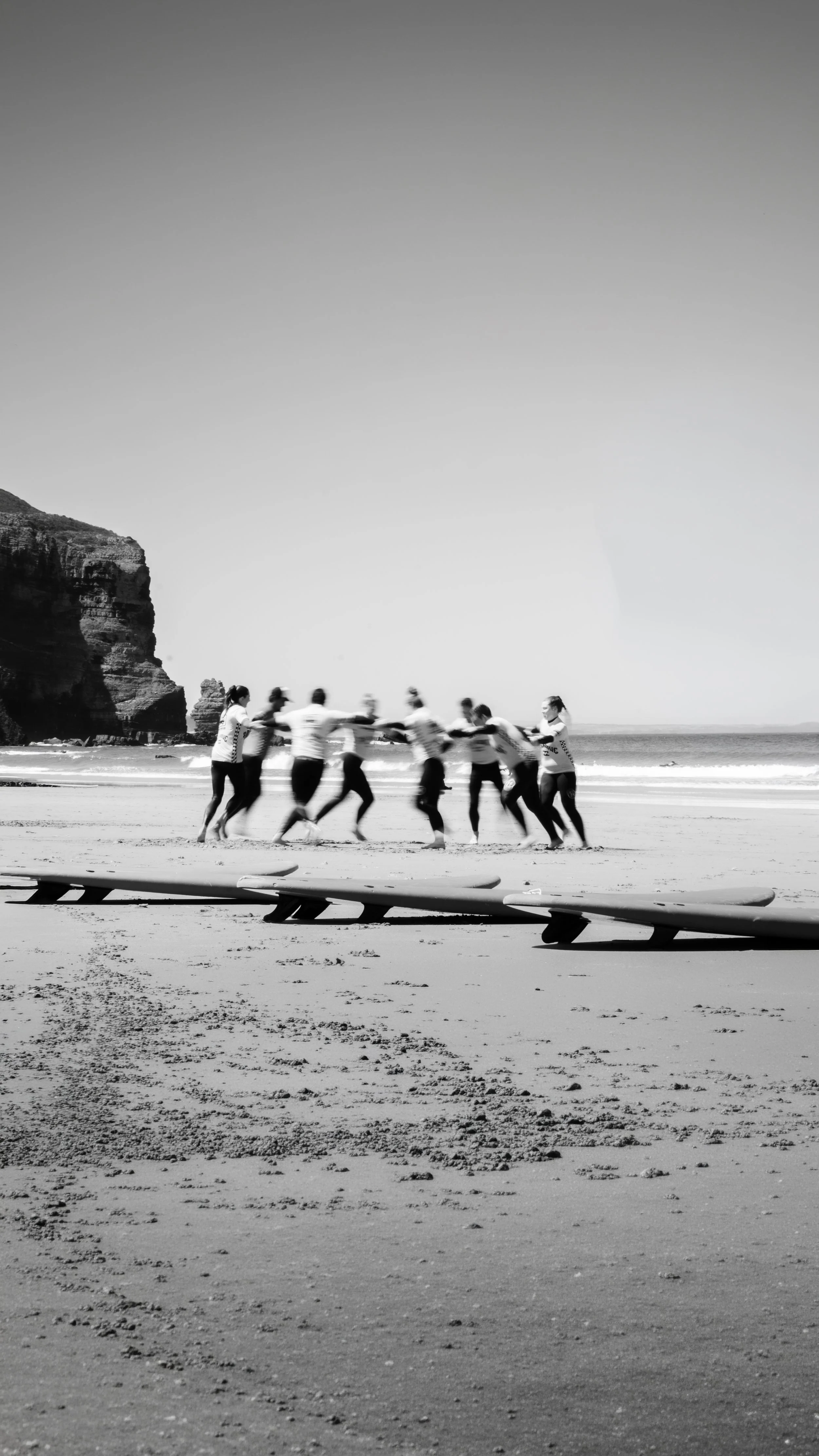 People playing tug-of-war on a beach with surfboards and cliffs in the background in black and white.