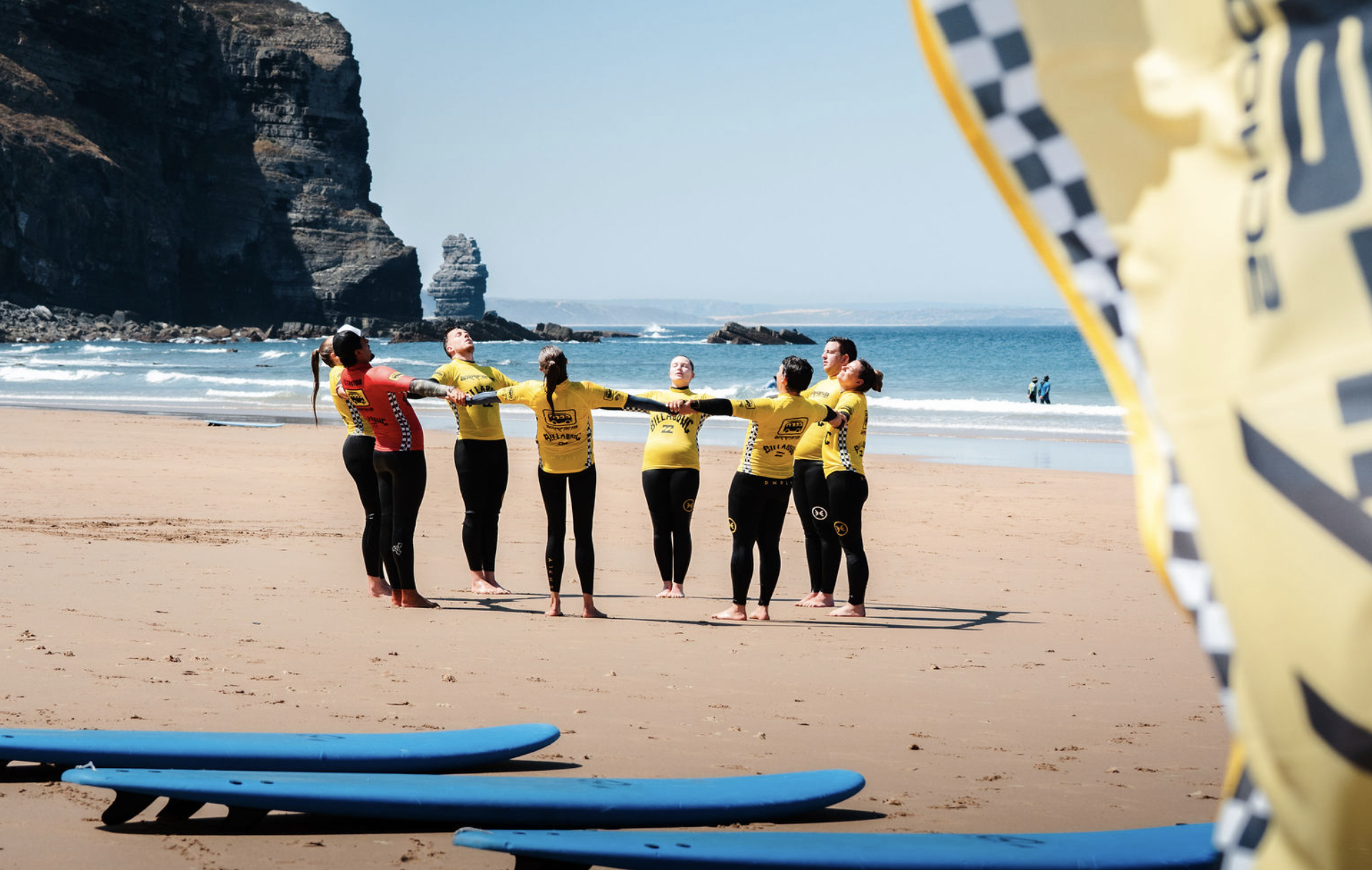 A group of people wearing wetsuits and surf gear standing on a sandy beach, holding hands in a circle, with surfboards lying on the sand nearby. The ocean and rocky cliffs are visible in the background.