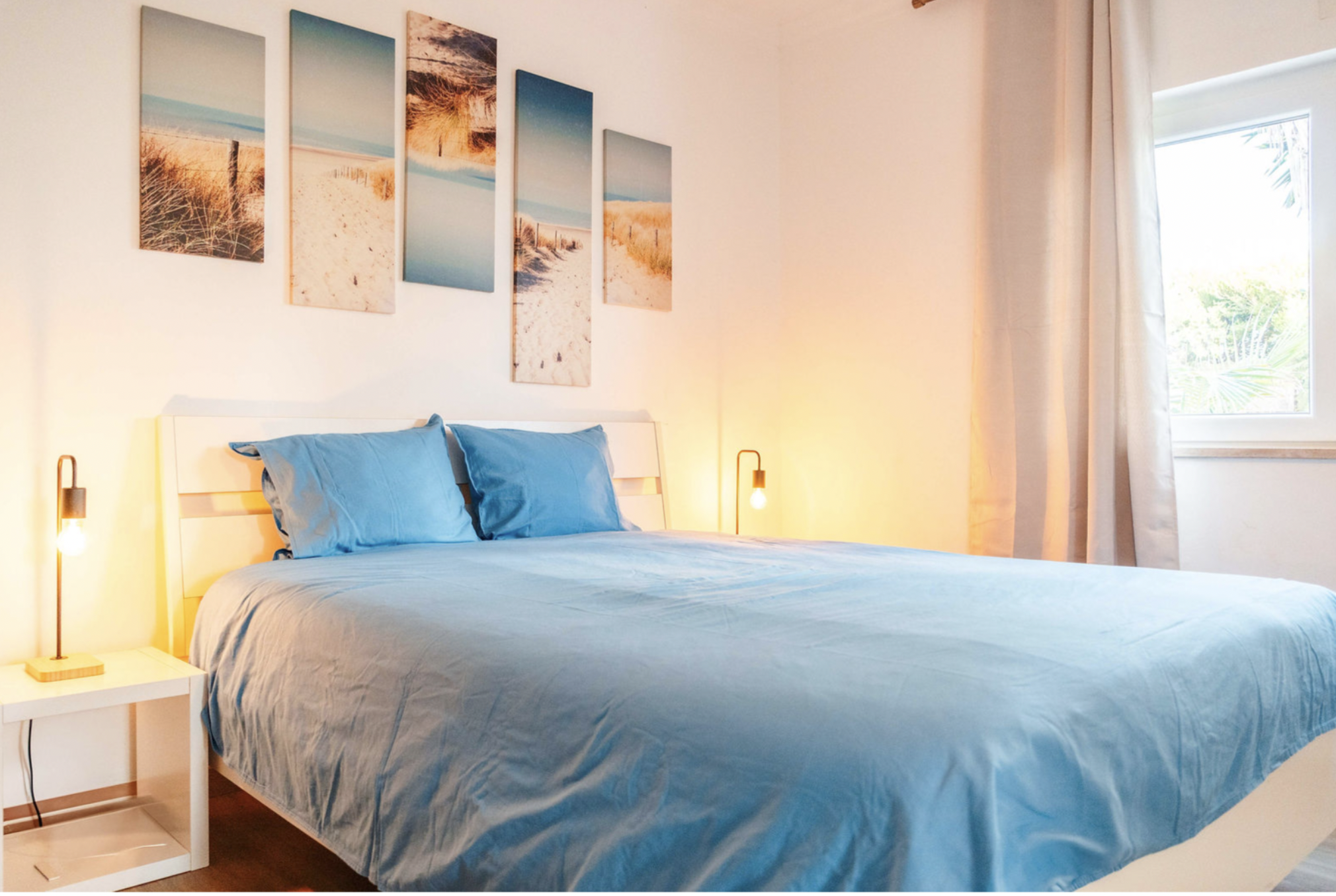 A bedroom with a bed covered in blue bedding and matching pillows, a white bedside table with a modern yellow lamp, a wall with five beach landscape paintings, an open window with white curtains, and a view of green foliage outside.