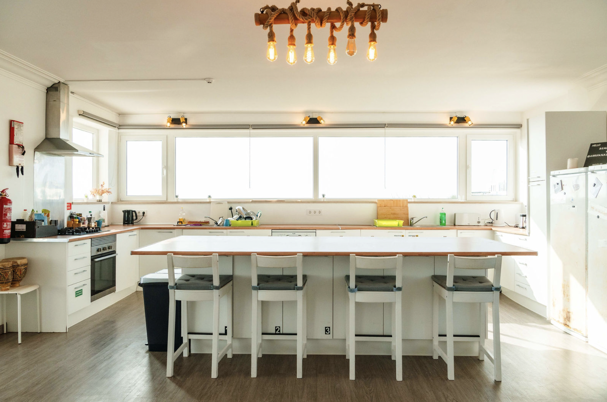 Modern kitchen with white cabinets, large windows, and a rectangular island with chairs. Overhead lighting includes a fixture with rope accents and Edison bulbs, and adjustable spotlights. Appliances such as a stainless steel oven, refrigerator, and 