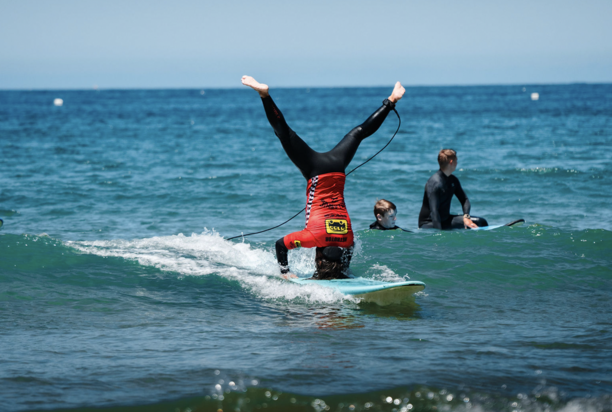 Person performing a handstand on a surfboard, balancing on the ocean, with two other individuals sitting on surfboards nearby, all wearing wetsuits