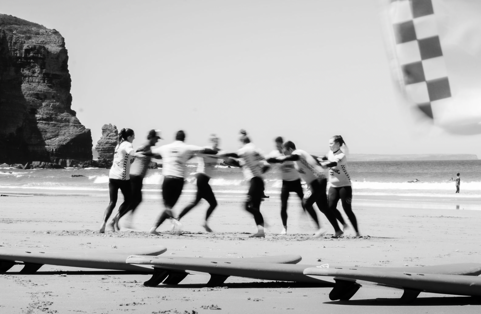 A group of people holding hands and playing on a beach with surfboards in the foreground, rocky cliffs on the left, a checkered sail or flag on the right, and the ocean in the background, all depicted in black and white.