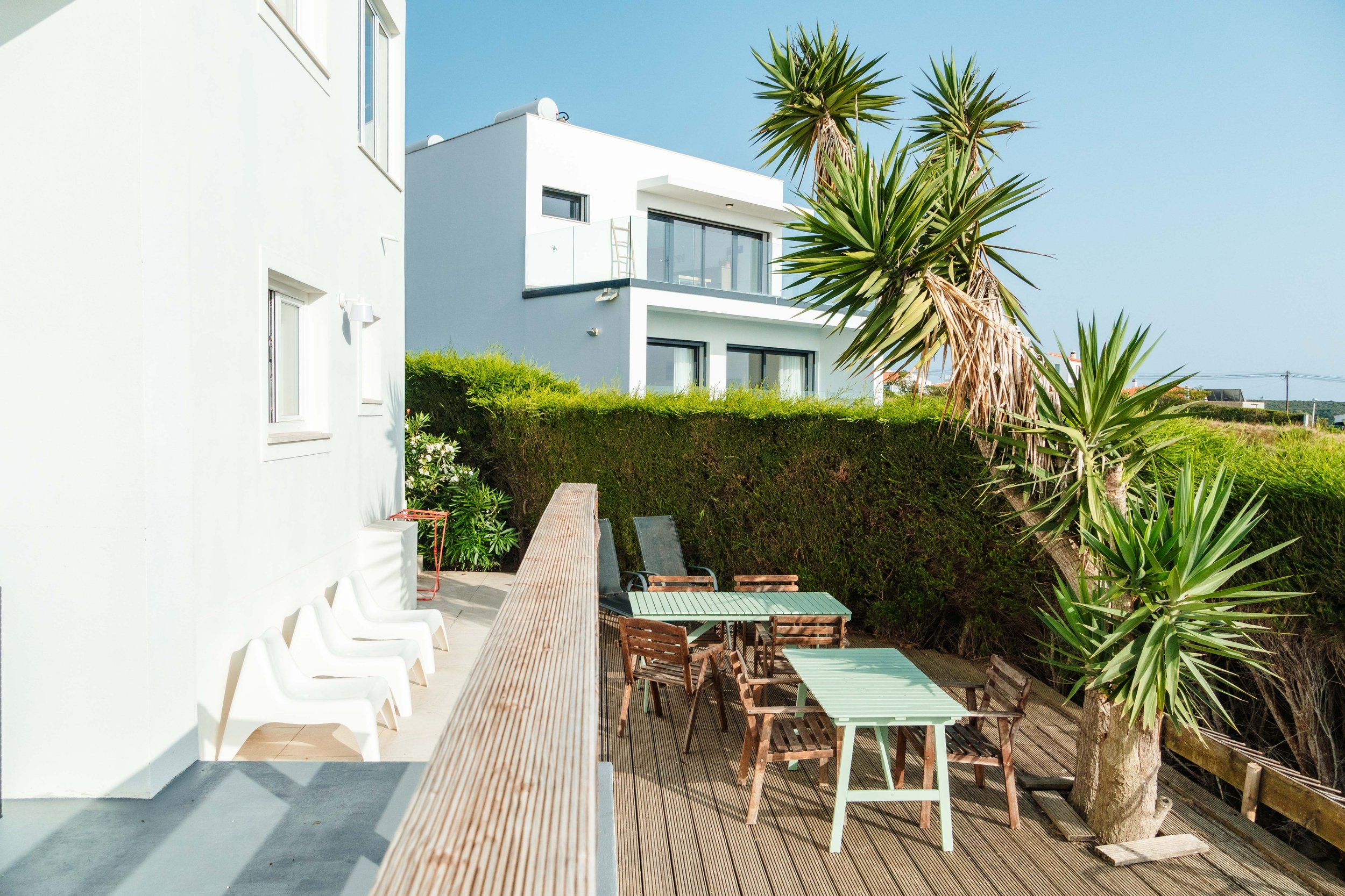 Outdoor patio with wooden tables and chairs, a row of white chairs against a white building, green bushes, and a tall palm tree under a clear blue sky.
