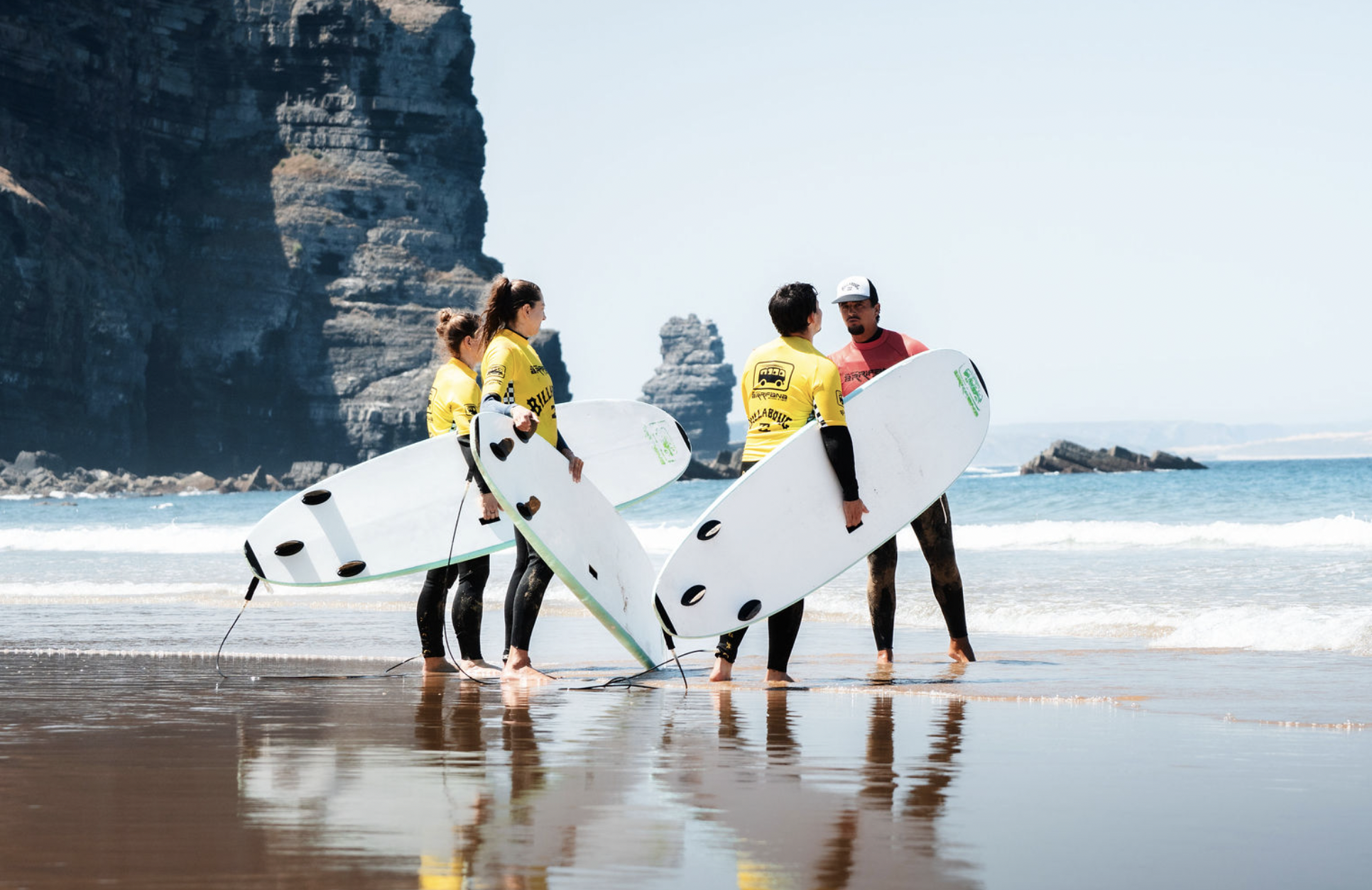 A group of four people at the beach with surfboards, wearing yellow and black wetsuits, standing in shallow water, with cliffs and rock formations in the background.