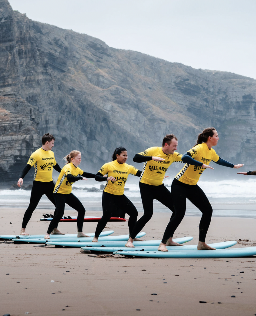 A group of five people in yellow shirts and black leggings are practicing surfing on a beach, standing on surfboards, with rocky cliffs and the ocean in the background.