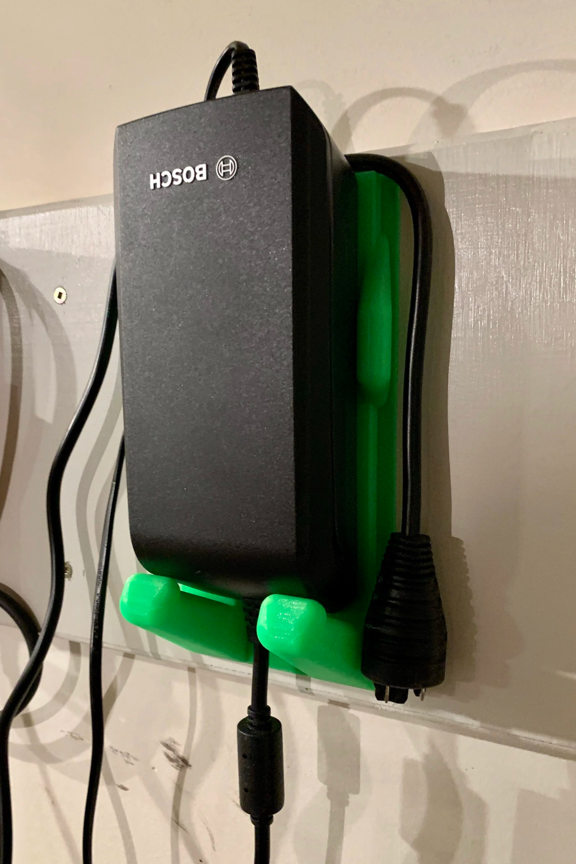 A Bosch electric e-bike charger mounted on a wall using a green plastic holder, with a power cord connected.