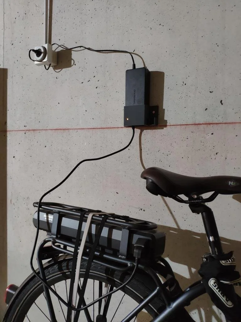 Electrical components attached to the wall, including a power outlet, a black box, and several cords connected to a bicycle with a mounted battery pack.