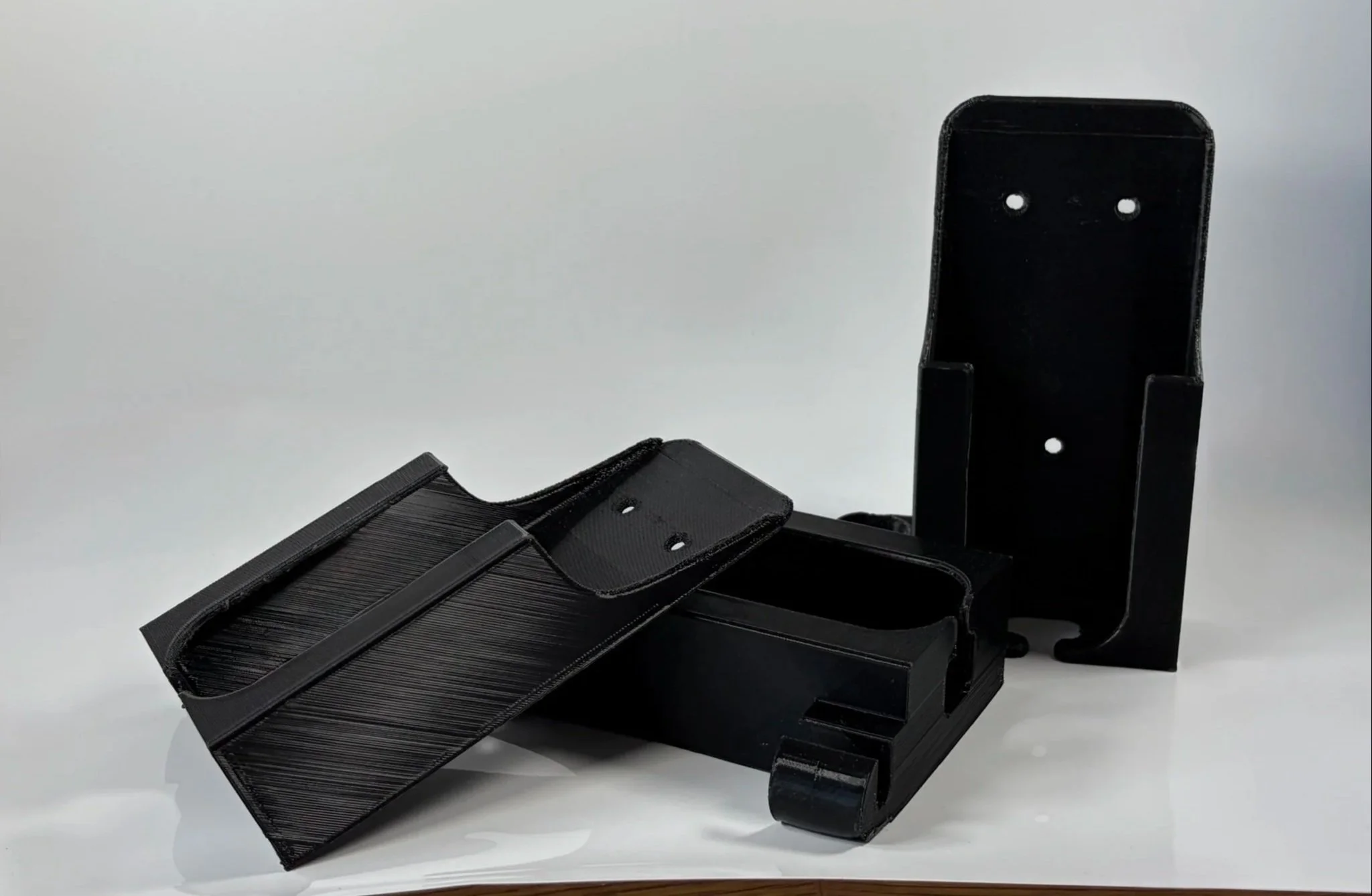 Three Black plastic E-Bike charger holders with two pieces, one lying flat and the other standing upright, on a white surface.
