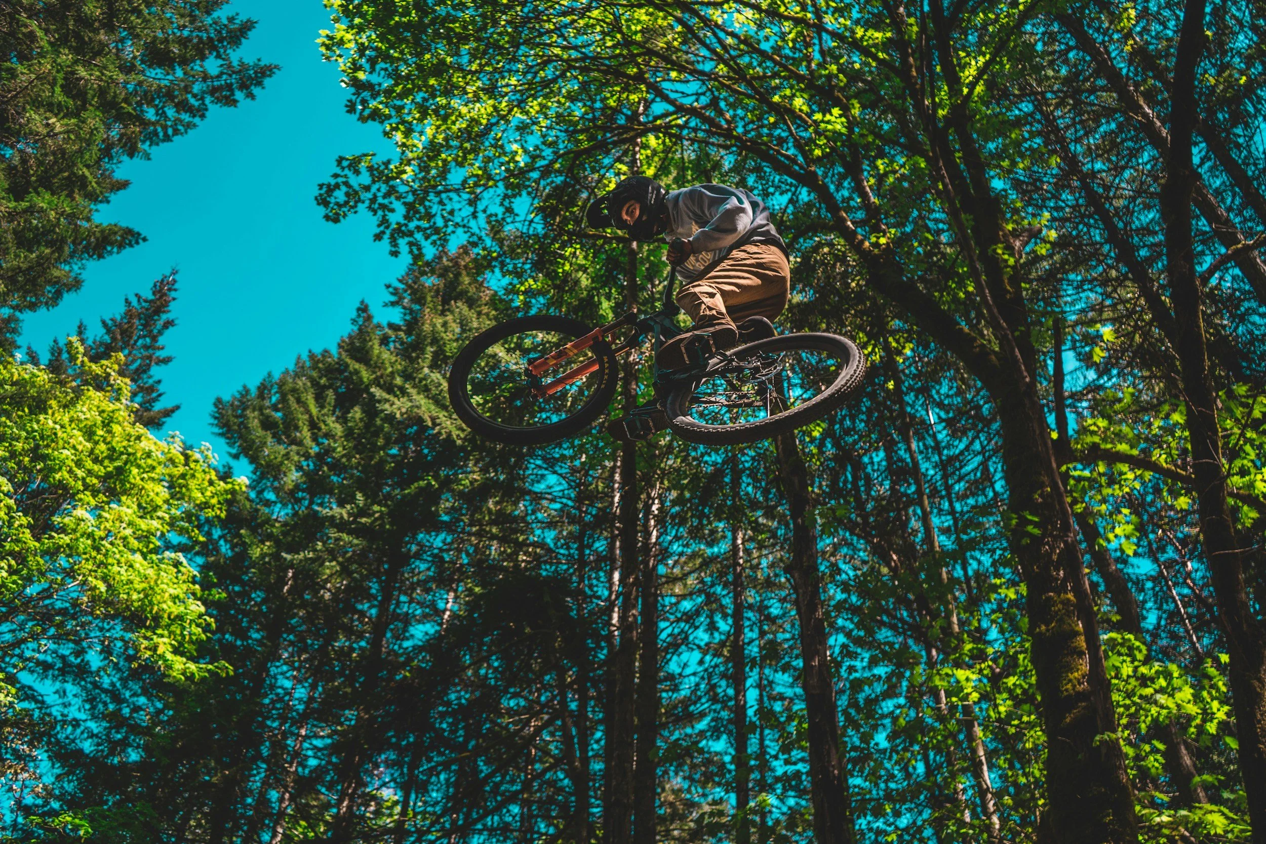 A person wearing a helmet and casual clothes performing an airborne stunt on a mountain bike, surrounded by tall trees with green foliage under a bright blue sky.