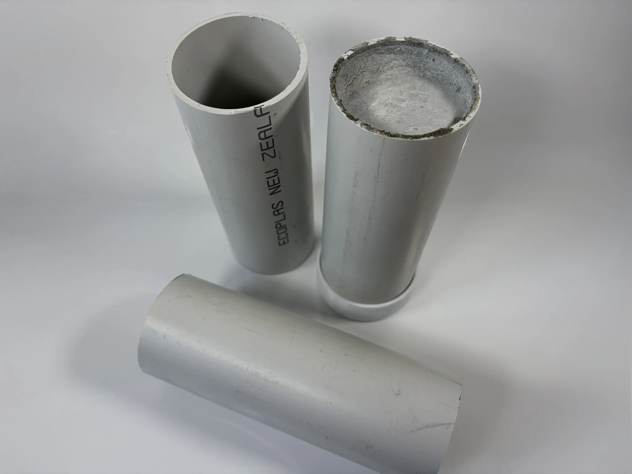 Three white PVC pipes, two standing upright and one lying horizontally, on a white surface. One pipe has markings 'ECOPLAS NEW ZEALAND'.