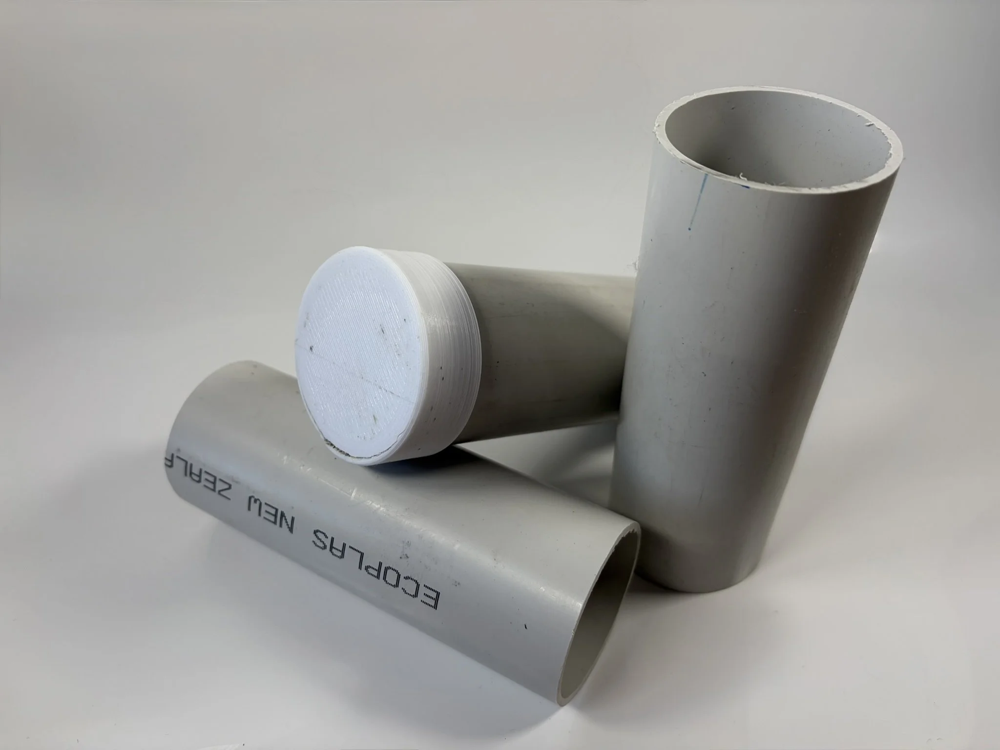 Three PVC pipe pieces on a white surface, one is a large cylindrical pipe, another is a smaller capped pipe, and the third is a medium-sized pipe with printing that reads 'ECOPLAS NEW ZEALAND'.