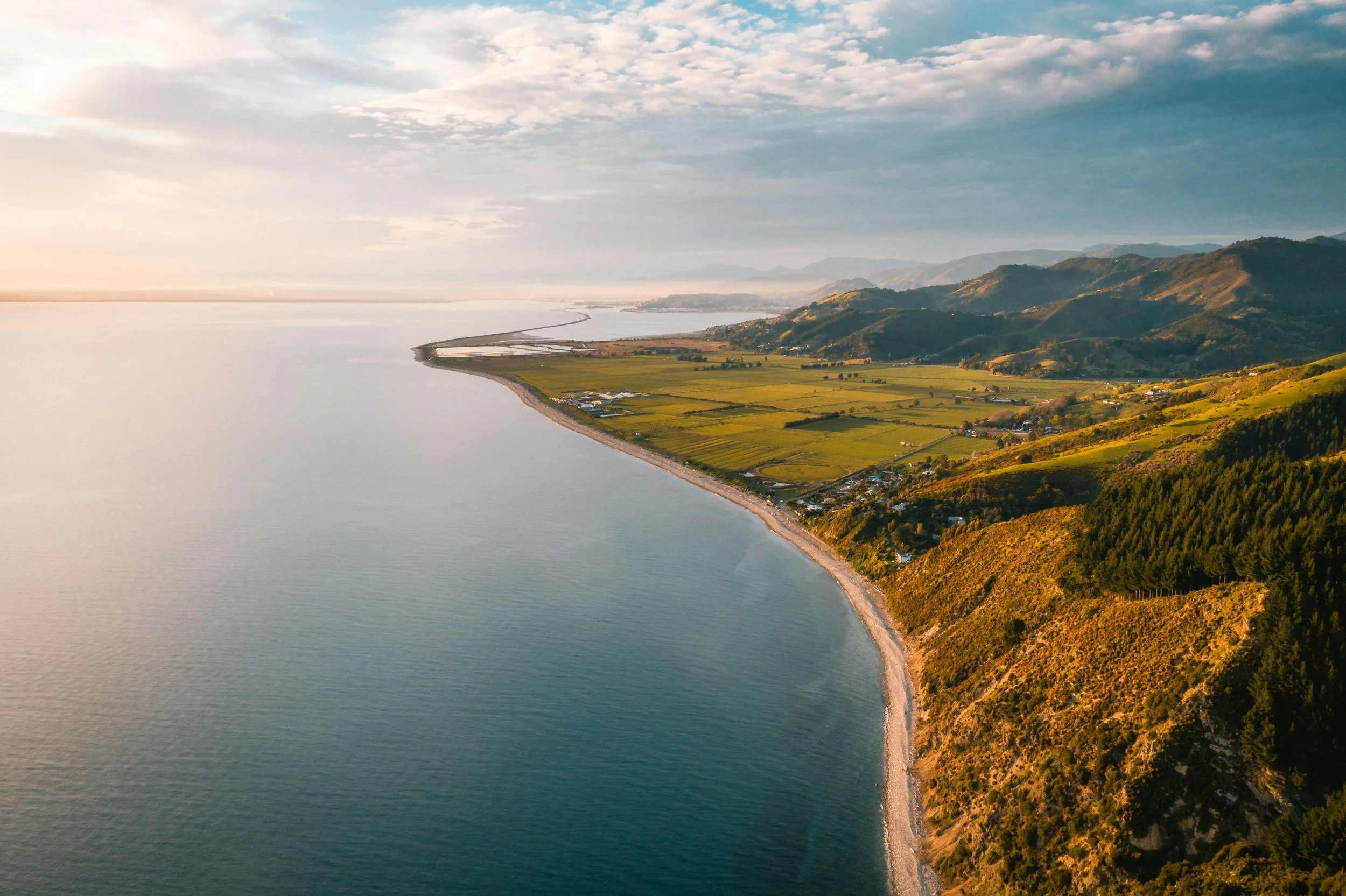 Aerial view of a coastal landscape with a beach, green fields, and hills at sunset. New Zealand.