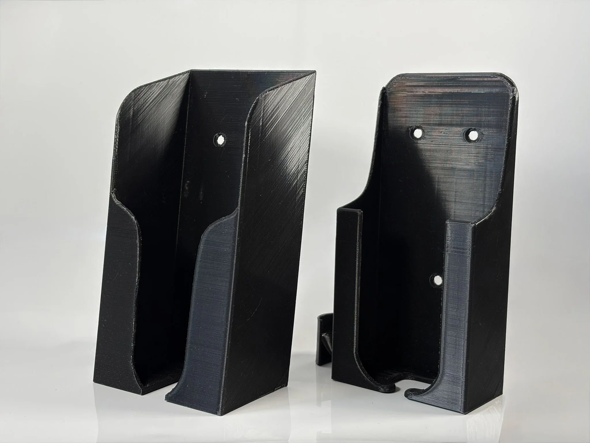 Two black 3D-printed wall mounts with holes for screws, showing different angles.